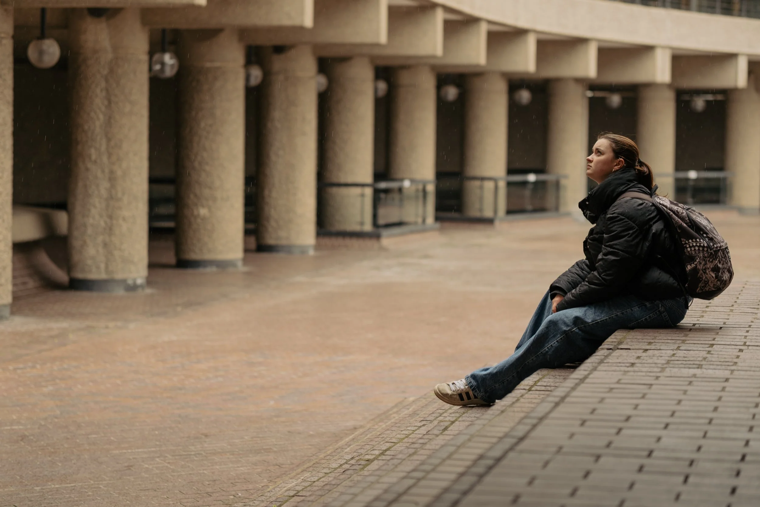 A woman sits alone on a city sidewalk near modern concrete columns, wearing a black jacket, jeans, and sneakers, with a backpack on her back, looking thoughtfully into the distance.