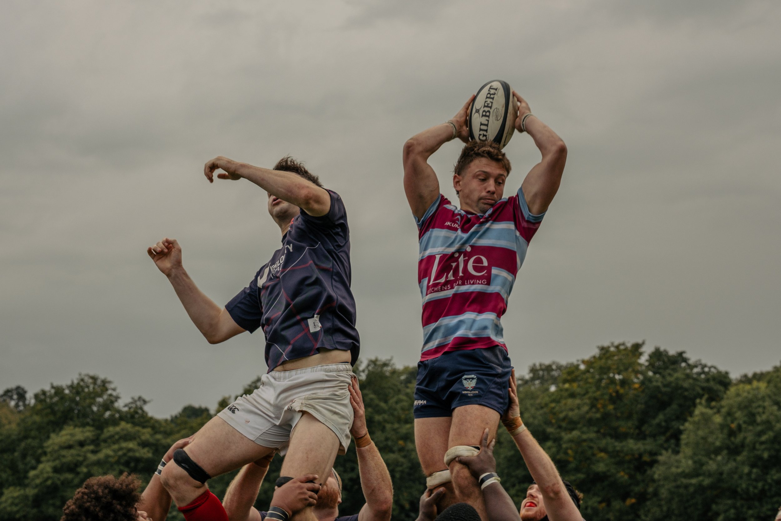 Two rugby players being lifted in a lineout during a match under a cloudy sky, with one player holding a rugby ball above his head.