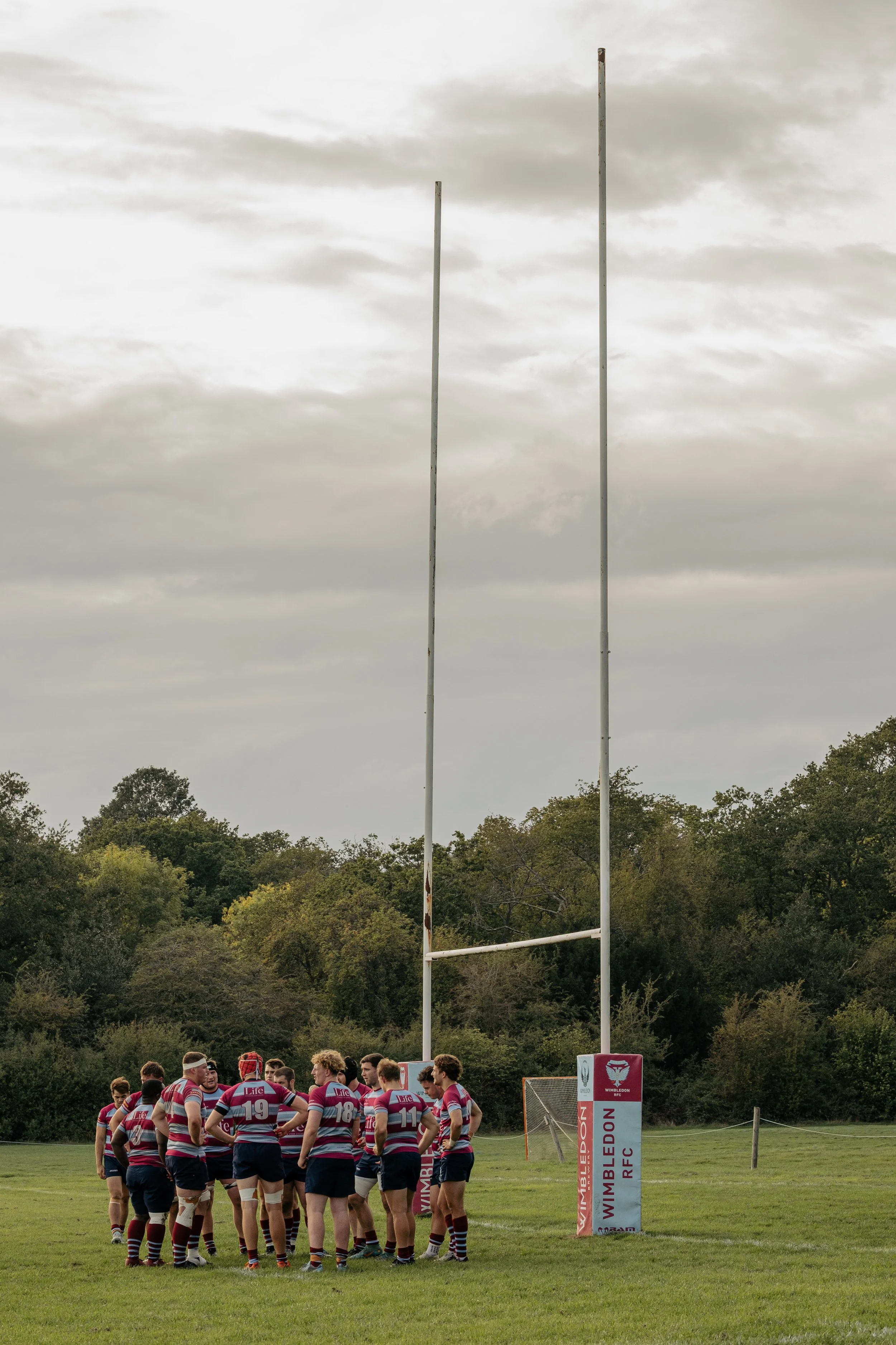 A rugby team gathered on the field during a game, with goalposts in the background and trees behind them.