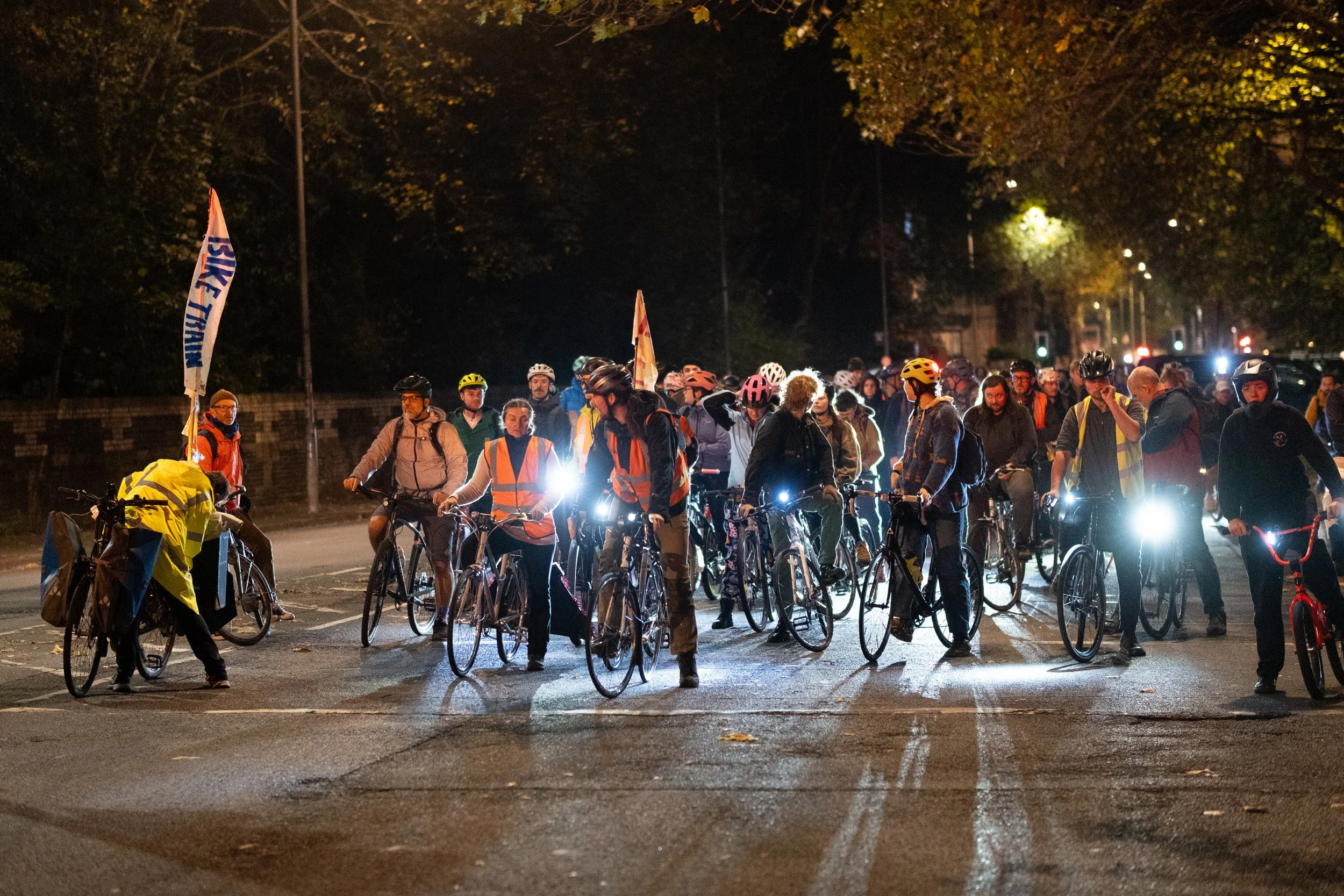 Group of cyclists gathered at night on a city street, some wearing helmets and reflective vests, with headlights illuminating the scene and trees lining the background.