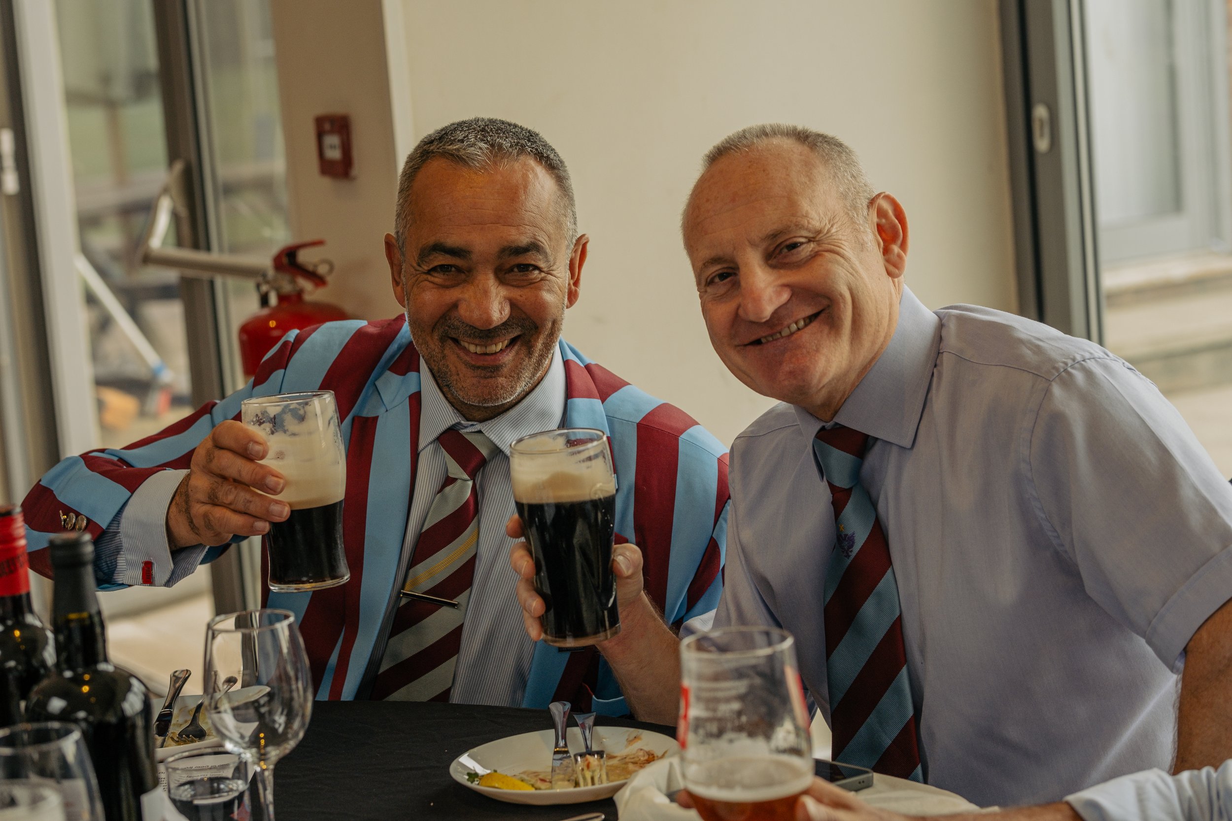 Two smiling men in dress shirts and ties holding glasses of dark beer with foam, sitting at a table with plates, glasses, and bottles, in a bright room with large windows.