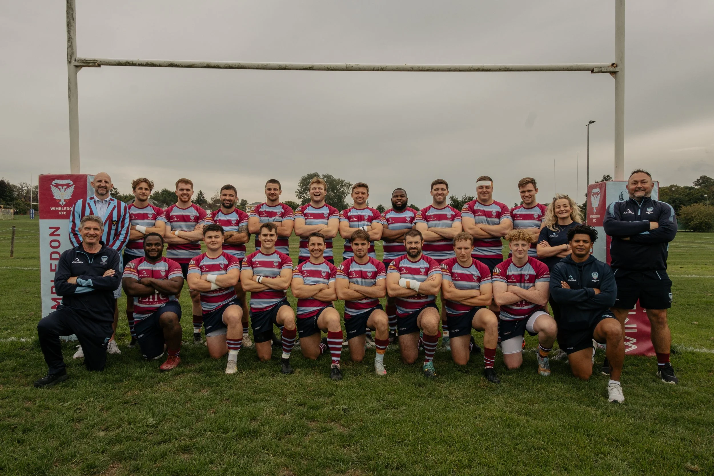 A rugby team and coaches posing for a group photo on a field with goal posts in the background. The team is wearing maroon and light blue striped jerseys, black shorts, and striped socks. The coaches are wearing dark sports jackets, some with the tea