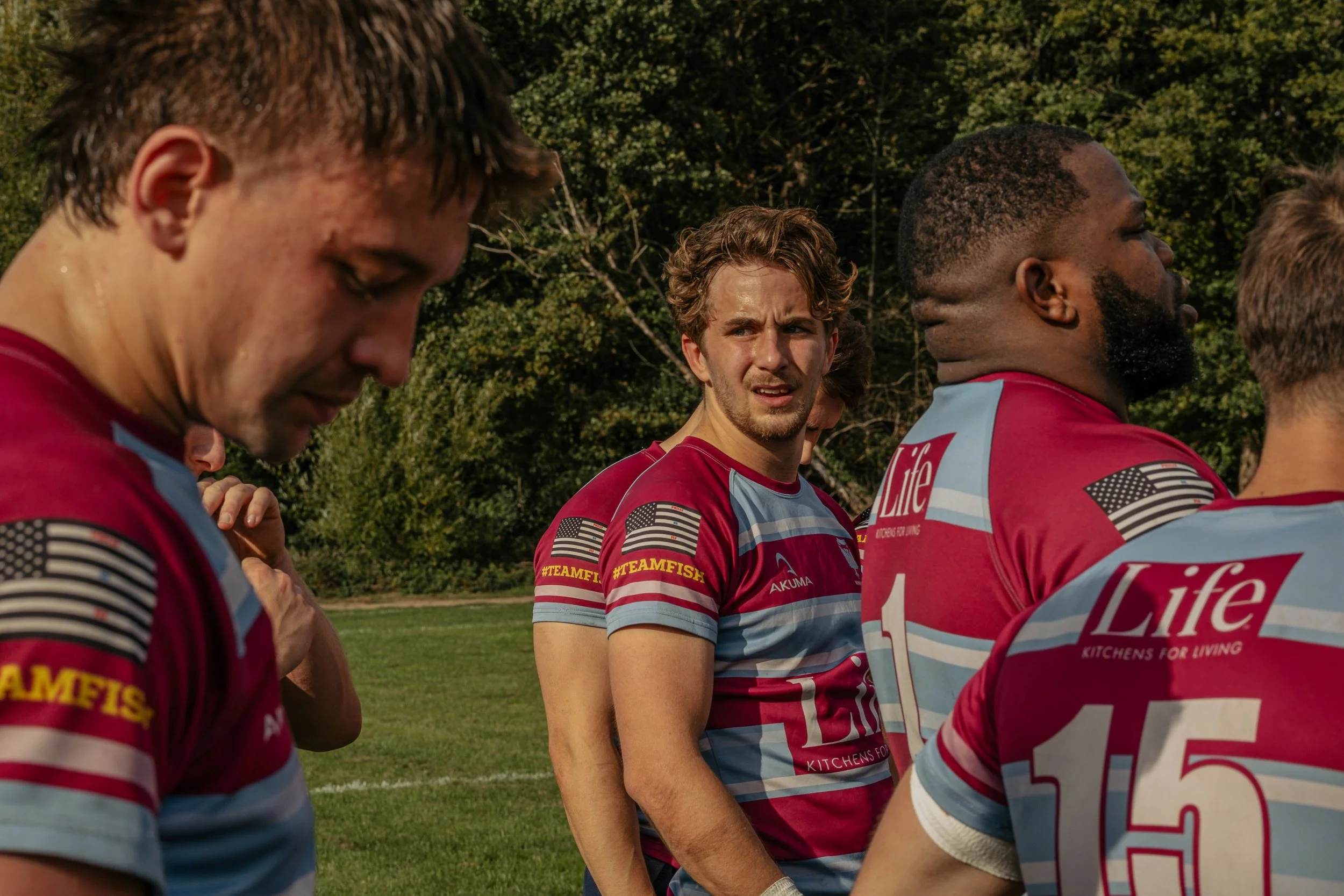 Group of young male rugby players in red and blue jerseys standing on a field, some with heads bowed, in a moment of reflection or prayer.