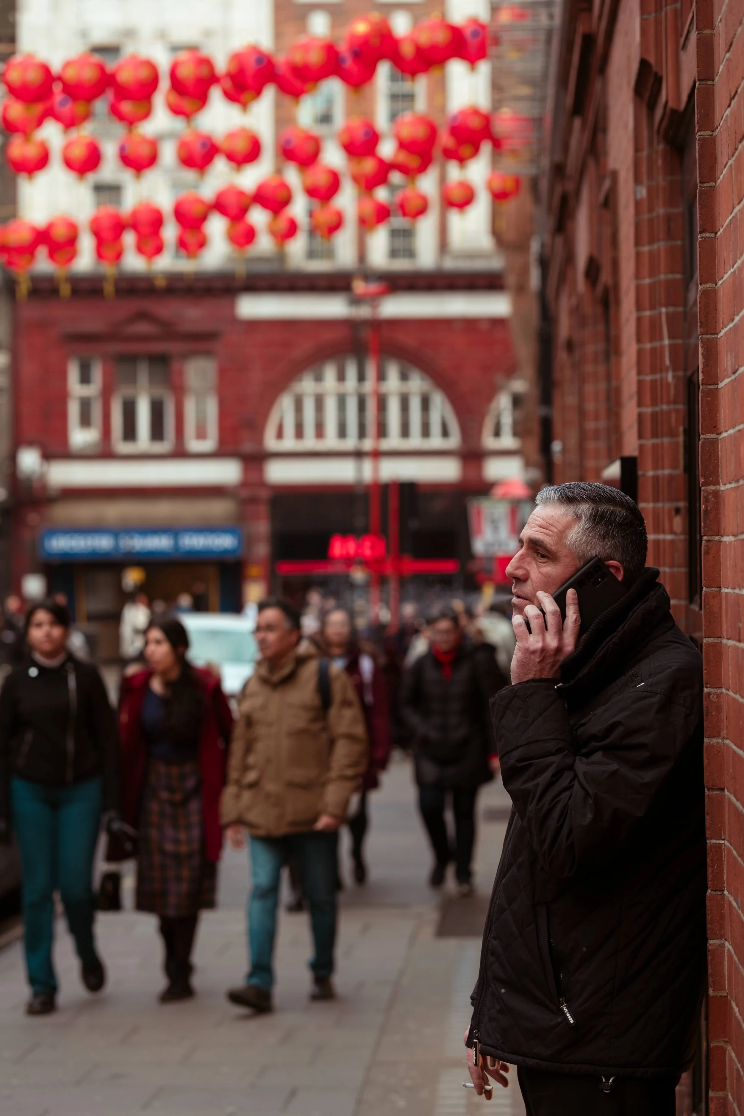 A man leaning against a brick wall talking on his cellphone in a busy urban street decorated with red Chinese lanterns.