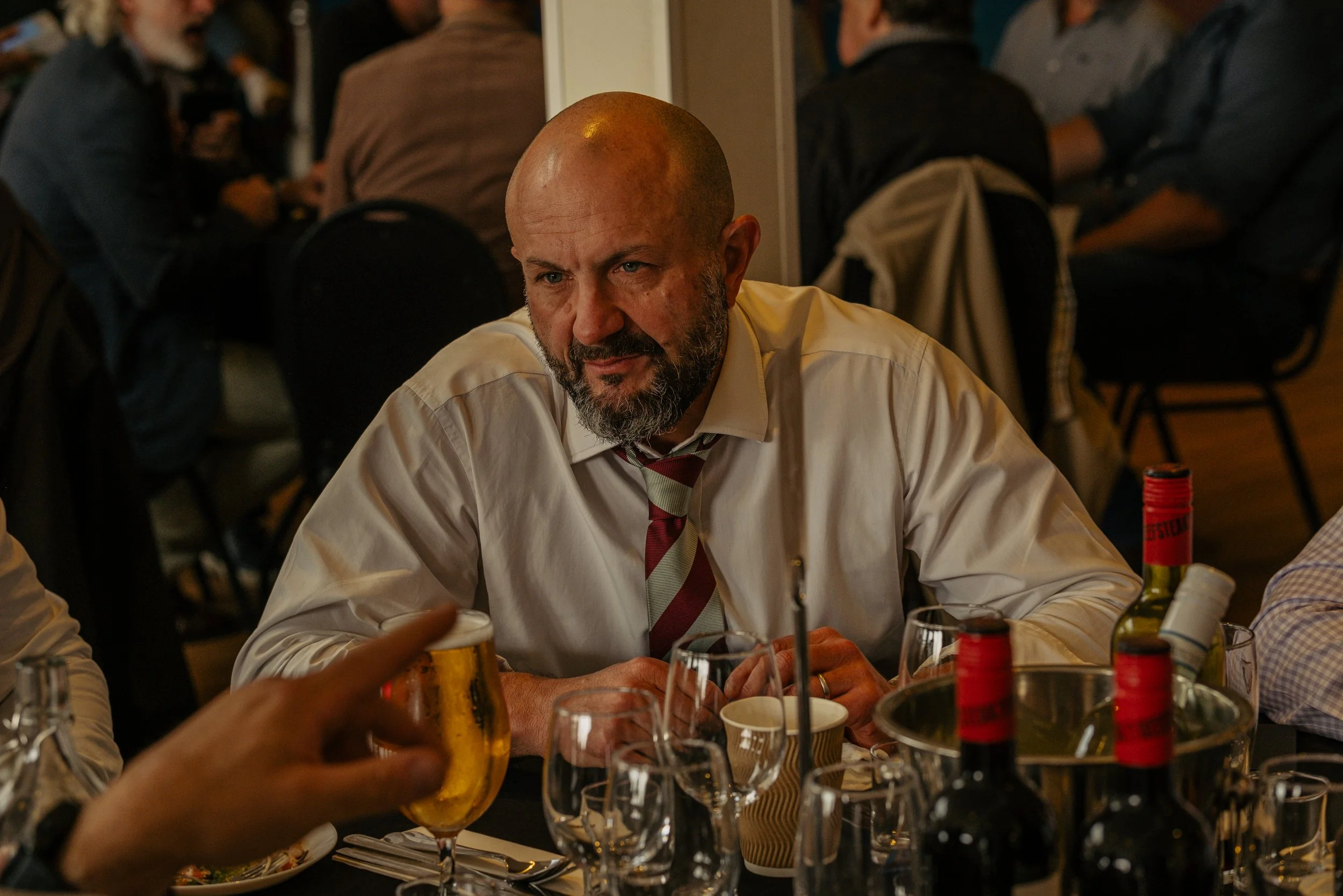 A man with a bald head and beard, wearing a white shirt and a red striped tie, sitting at a table with drinks in a restaurant or bar, with several wine bottles and glasses on the table. The background shows other people sitting and socializing.