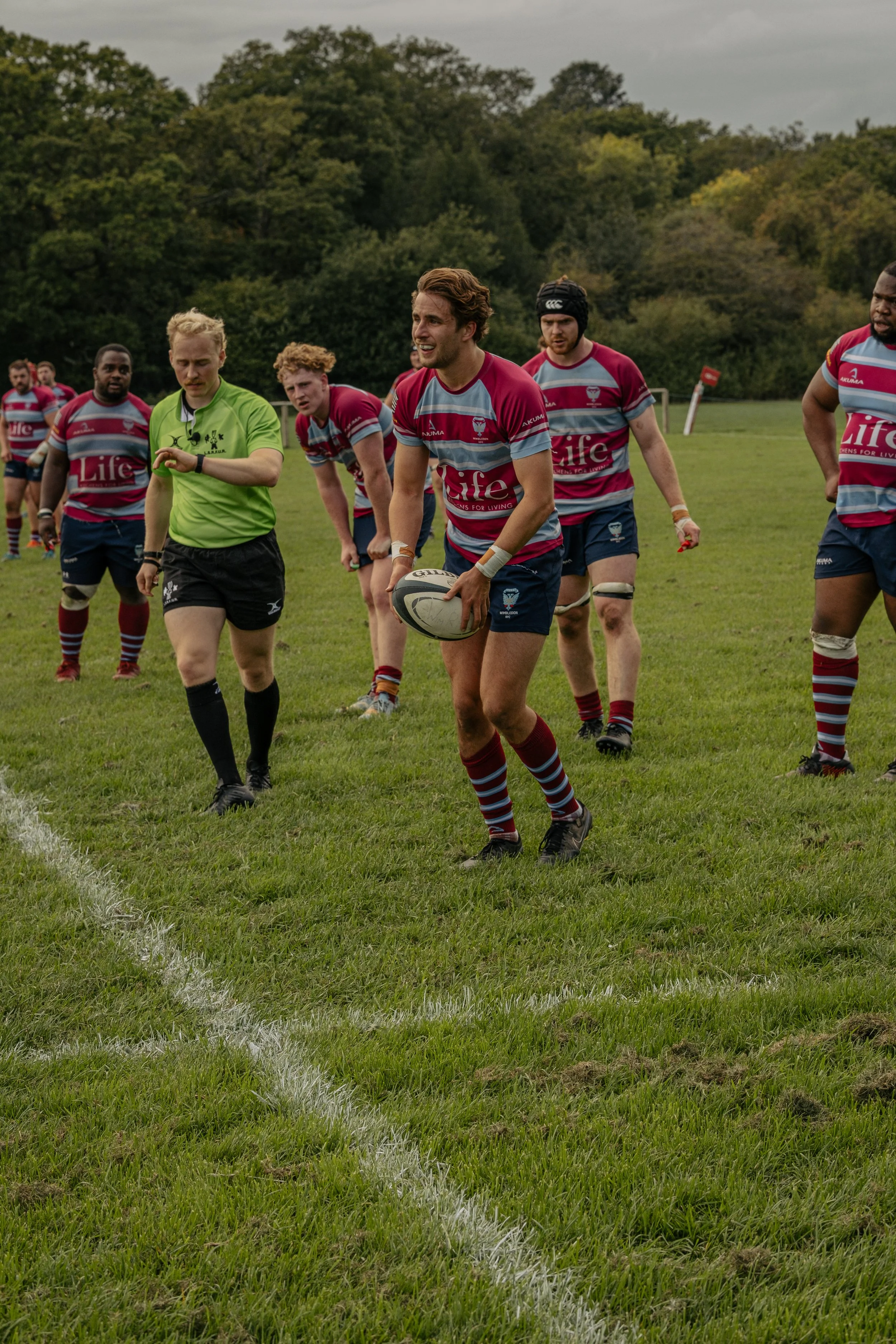 Rugby players walking back after a game, with one player holding a rugby ball, on a grassy field surrounded by trees.