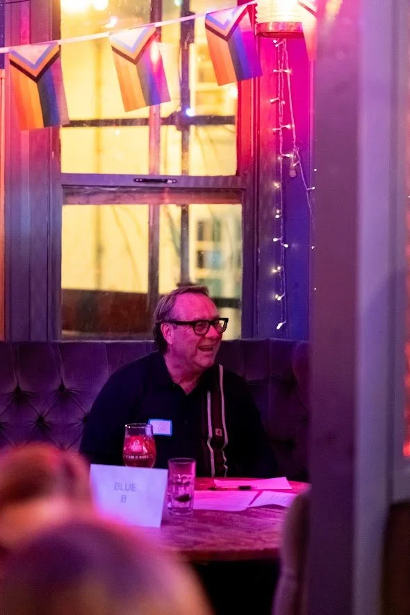 A man sitting at a dining table, smiling, in a decorated room with colorful flags and string lights, during a celebration.