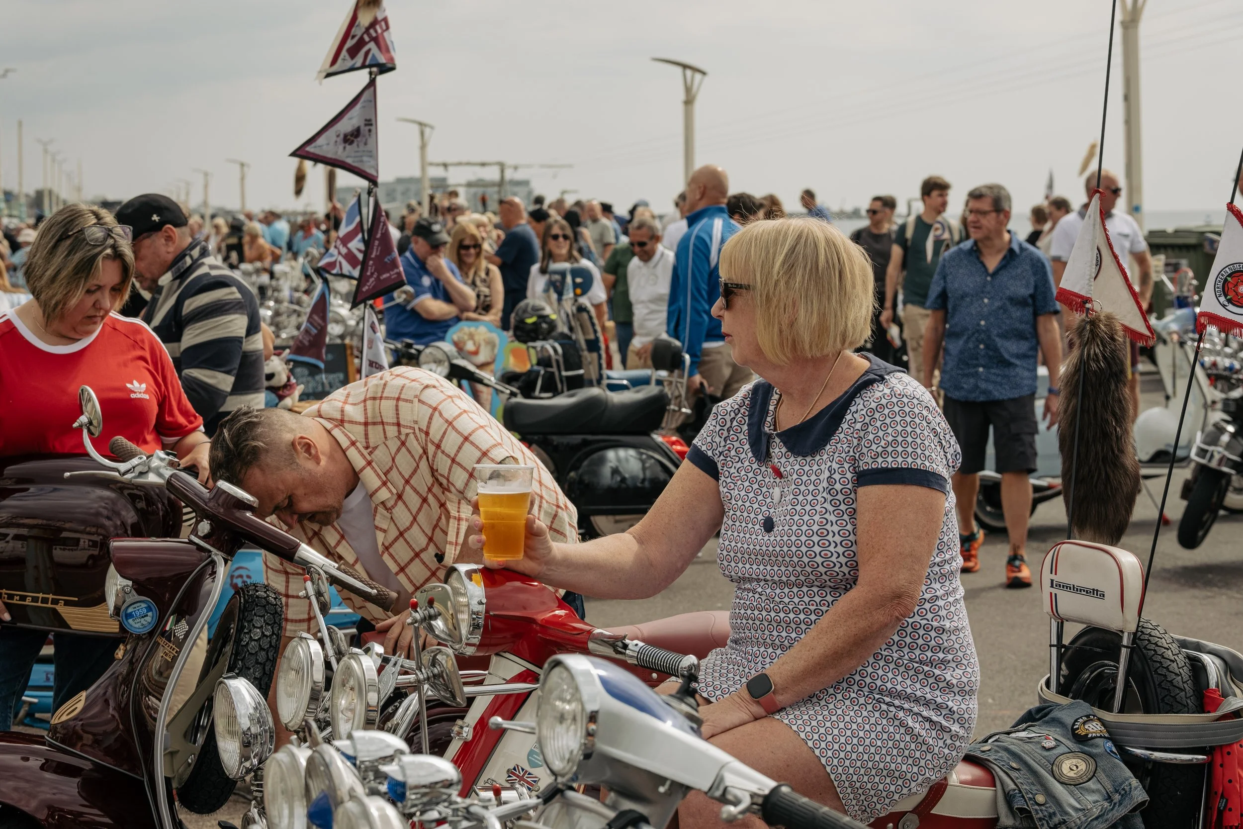 People attending a vintage motorcycle event, with motorcycles on display and flags, in an outdoor setting on a cloudy day.