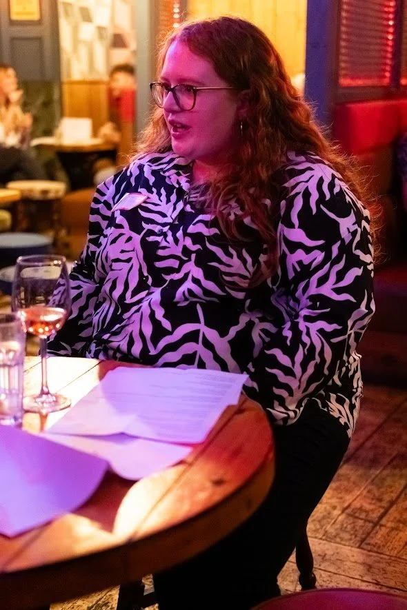 A woman with long curly hair, glasses, and a zebra print shirt sitting at a wooden table in a restaurant, with glasses and papers on the table, and other people dining in the background.