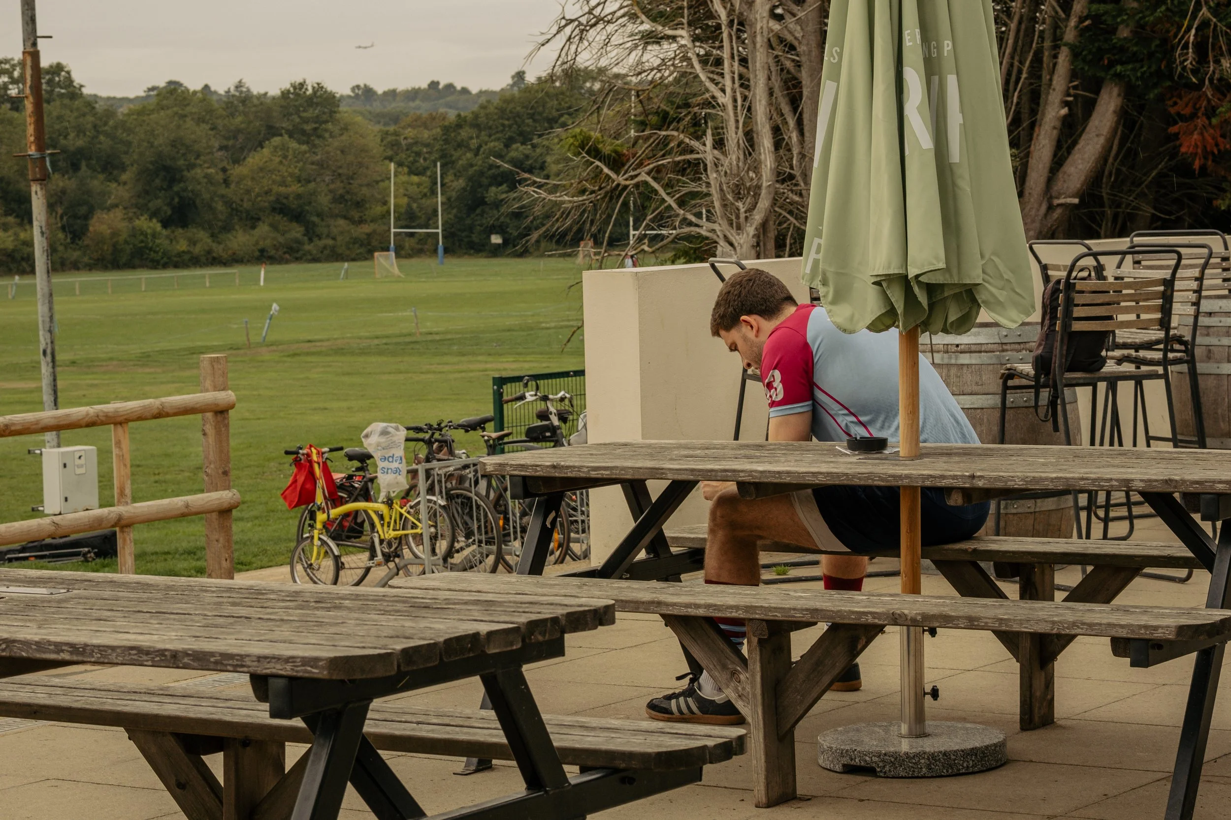 A young man sits alone outdoors at a picnic table with his head down, near a sports field with bicycles parked nearby, under a large green umbrella.
