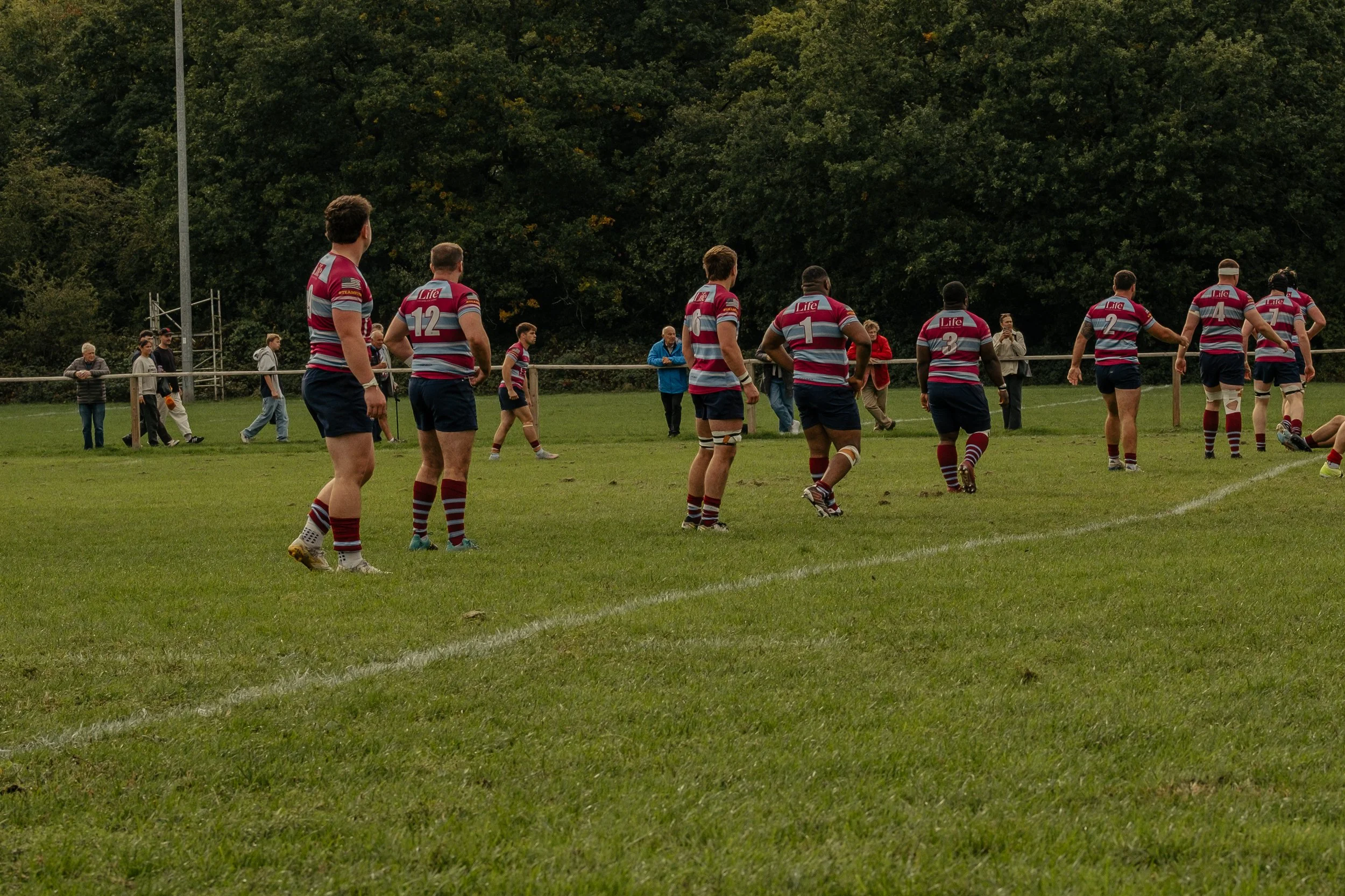 Rugby players in striped uniforms warming up on a grassy field with spectators watching from the sidelines.