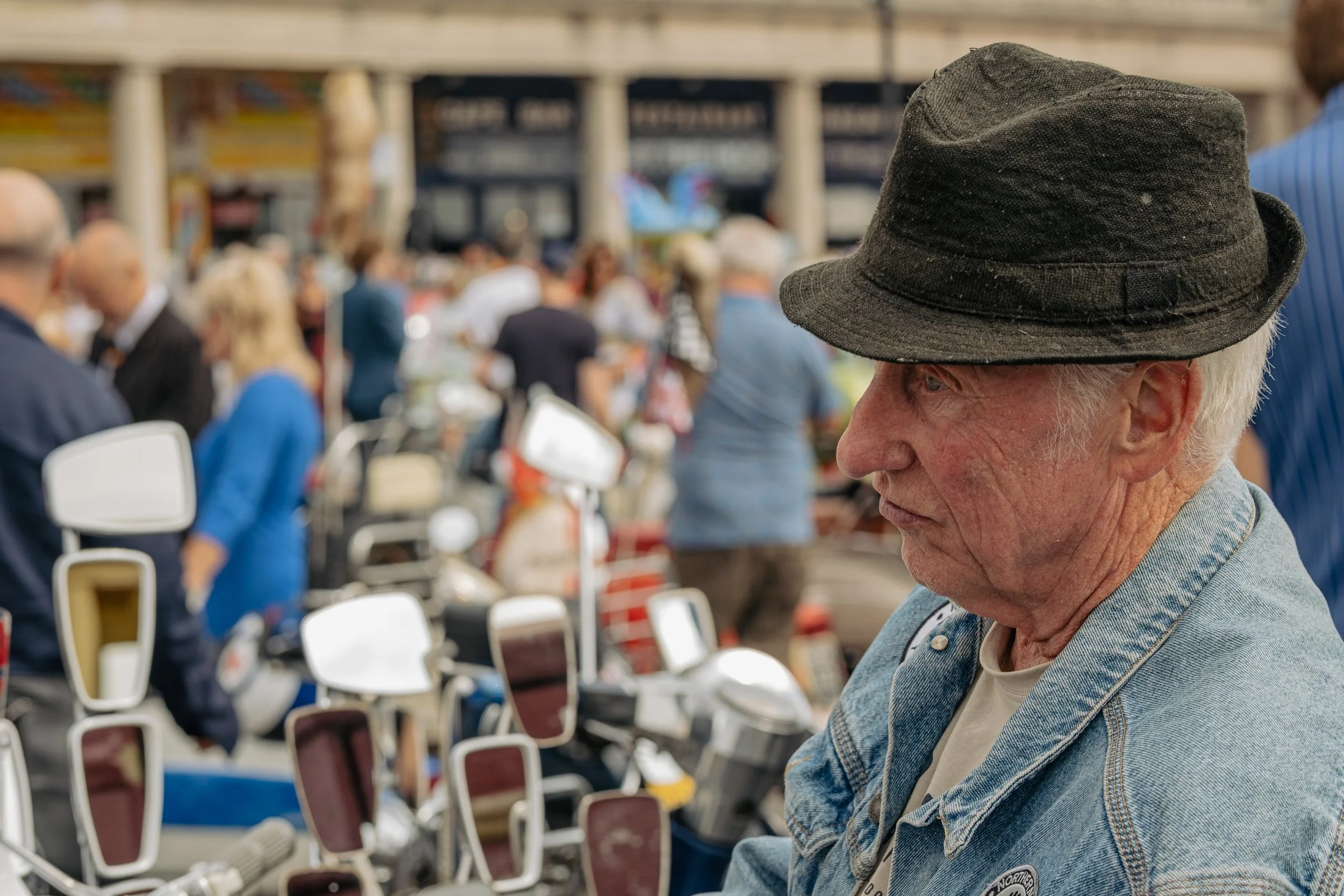 An elderly man wearing a black hat and denim jacket, with motorcycles and a crowd of people in the background at an outdoor event.