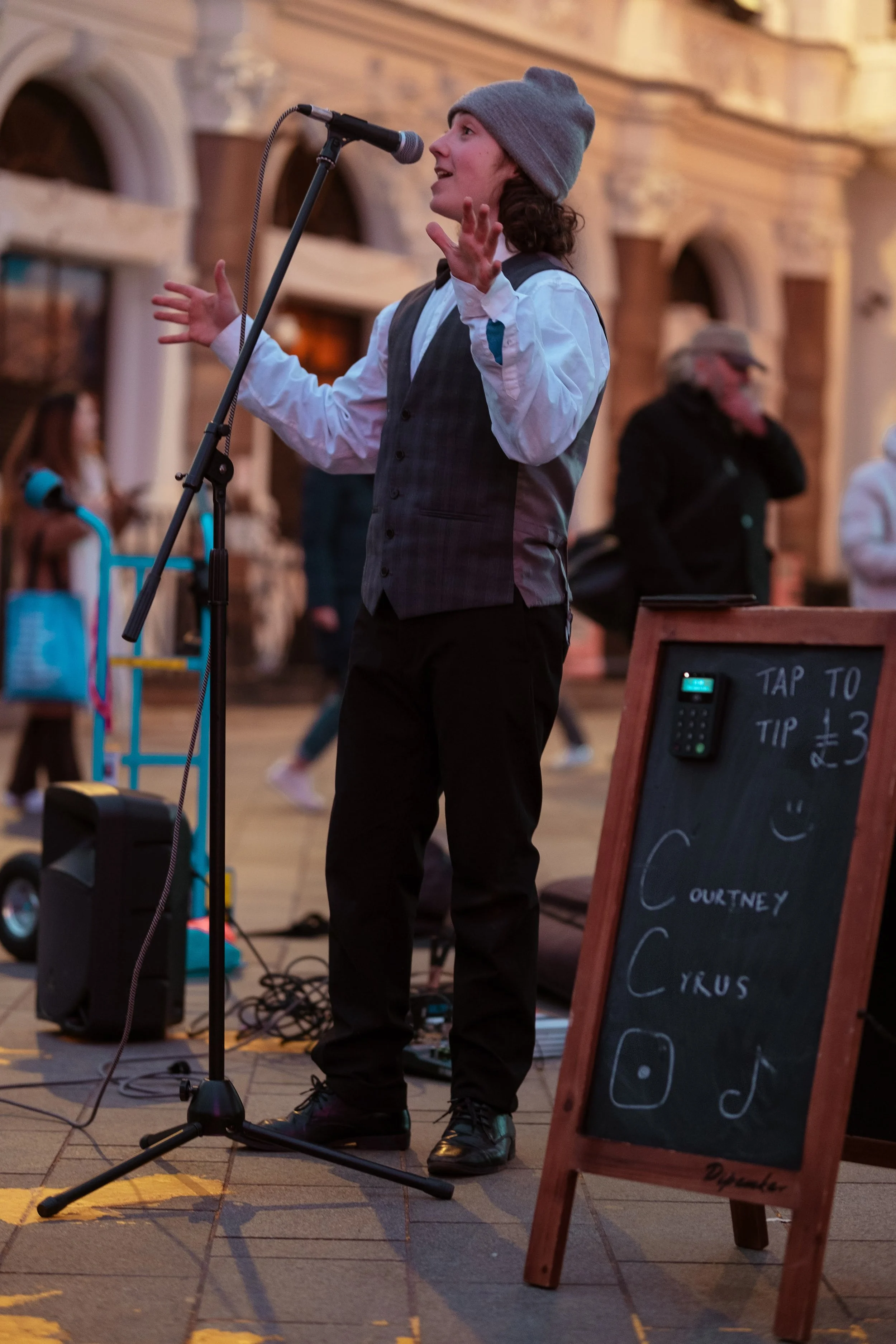 A street performer, a young man in a gray beanie, white shirt, and vest, singing into a microphone with his hands raised, standing on a city street with a crowd in the background. There is a blackboard sign nearby advertising tips and social media ic