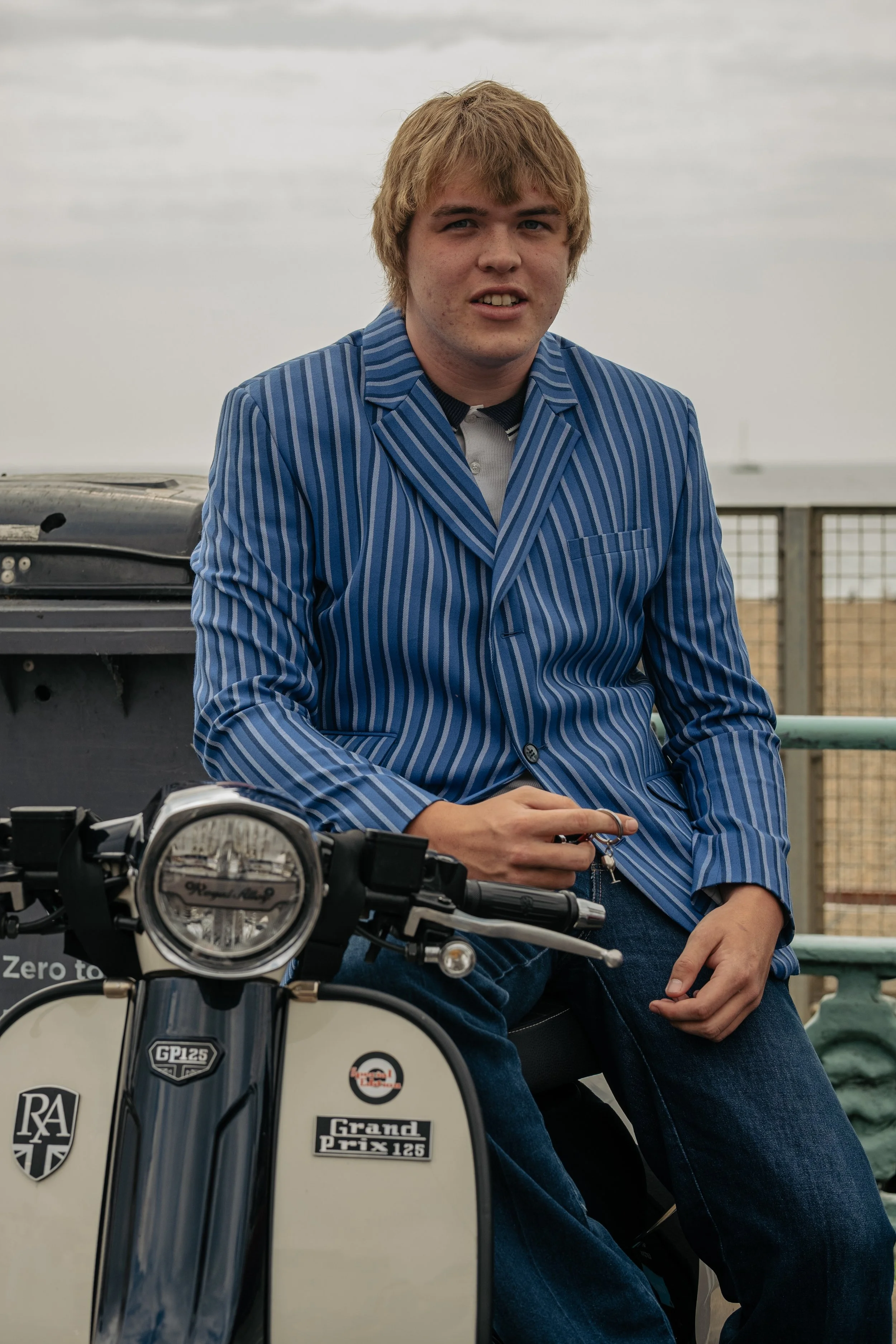 A young man with light brown hair wearing a blue striped blazer sitting on a vintage motorcycle near a railing, with a cloudy sky in the background.