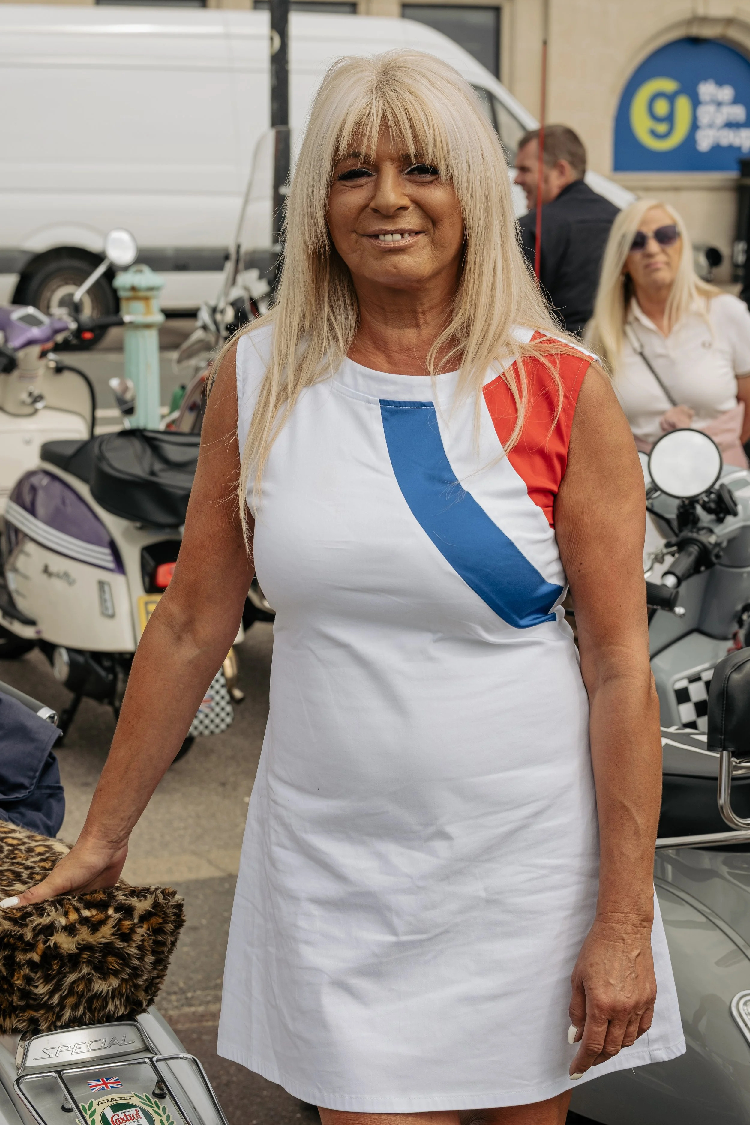 A woman with long blonde hair standing outdoors among scooters and motorcycles, smiling, wearing a white dress with red and blue accents.