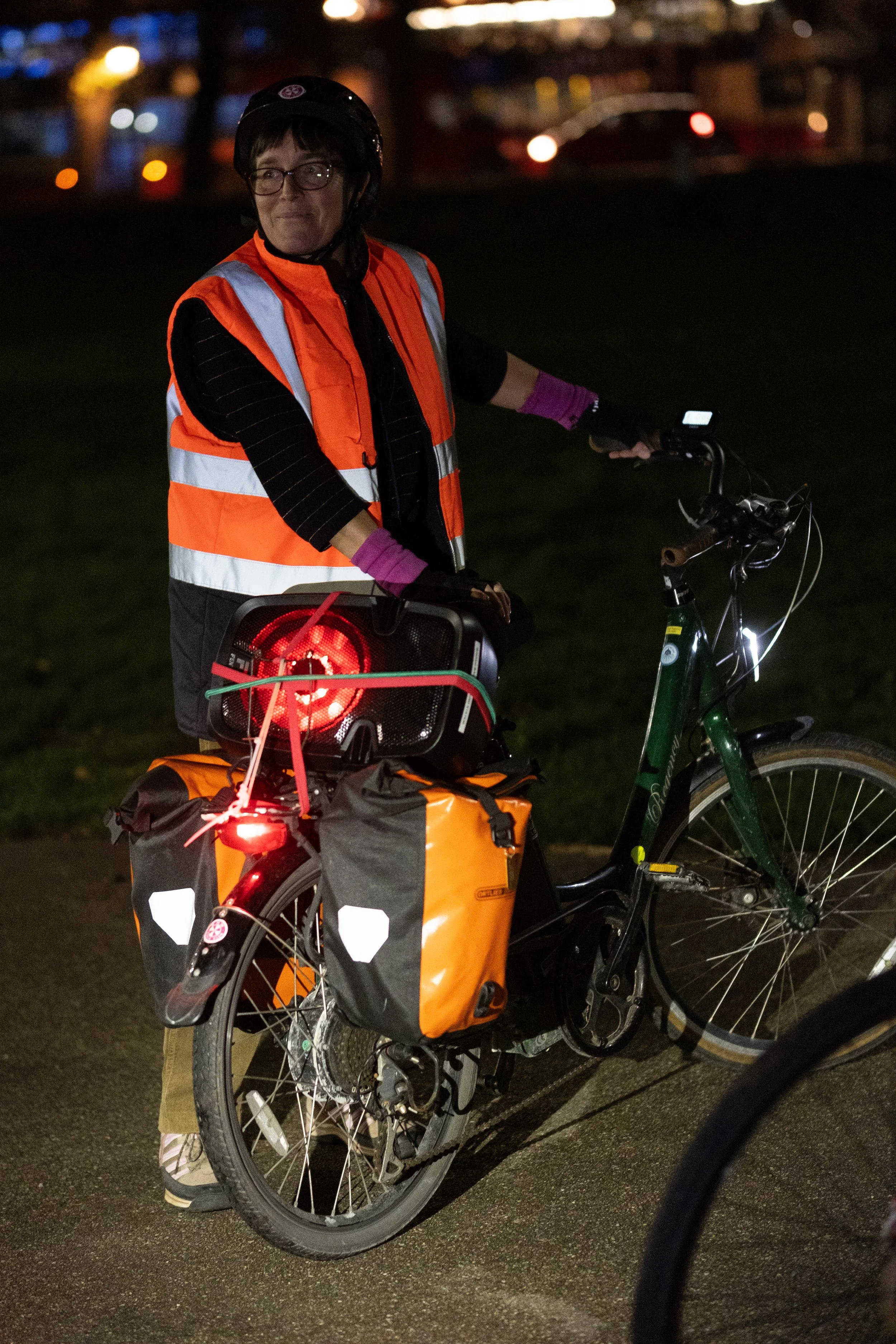 A woman wearing a helmet, glasses, a black striped shirt, an orange reflective safety vest, purple gloves, and gray pants standing outdoors at night with a bicycle equipped with orange panniers and a lit rear light.