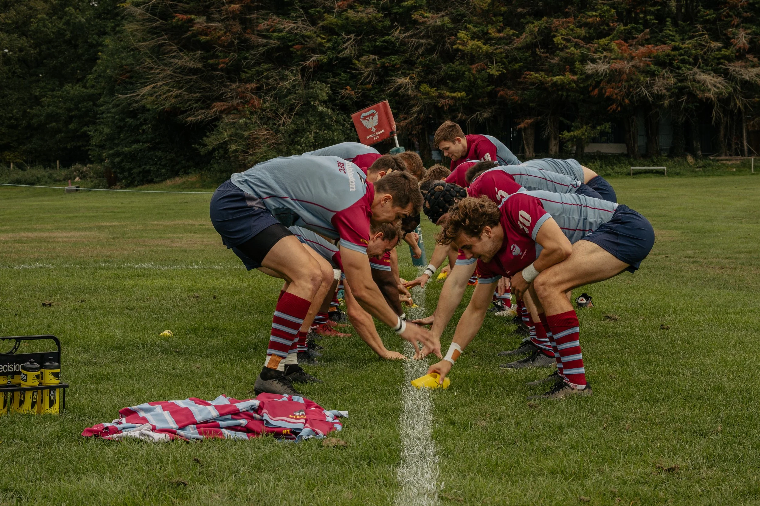 A rugby team is preparing on the field, with players kneeling and pouring water over the white line on the grass.