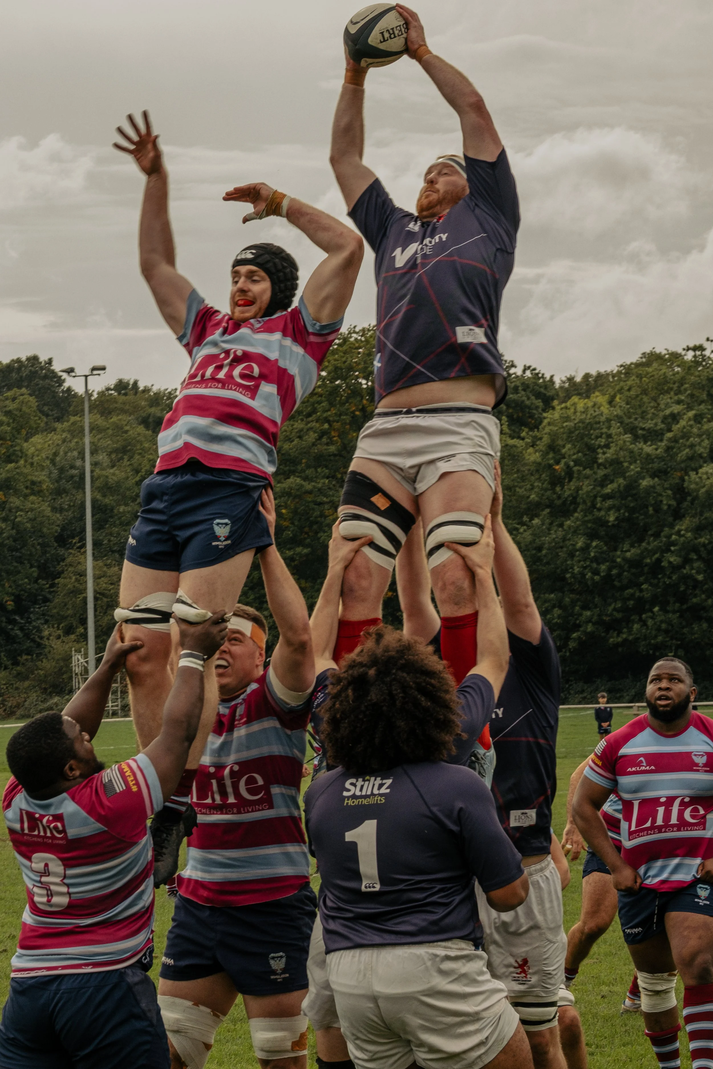 Rugby players in a lineout, with one player lifting another to catch the ball, on a rugby field with trees in the background.