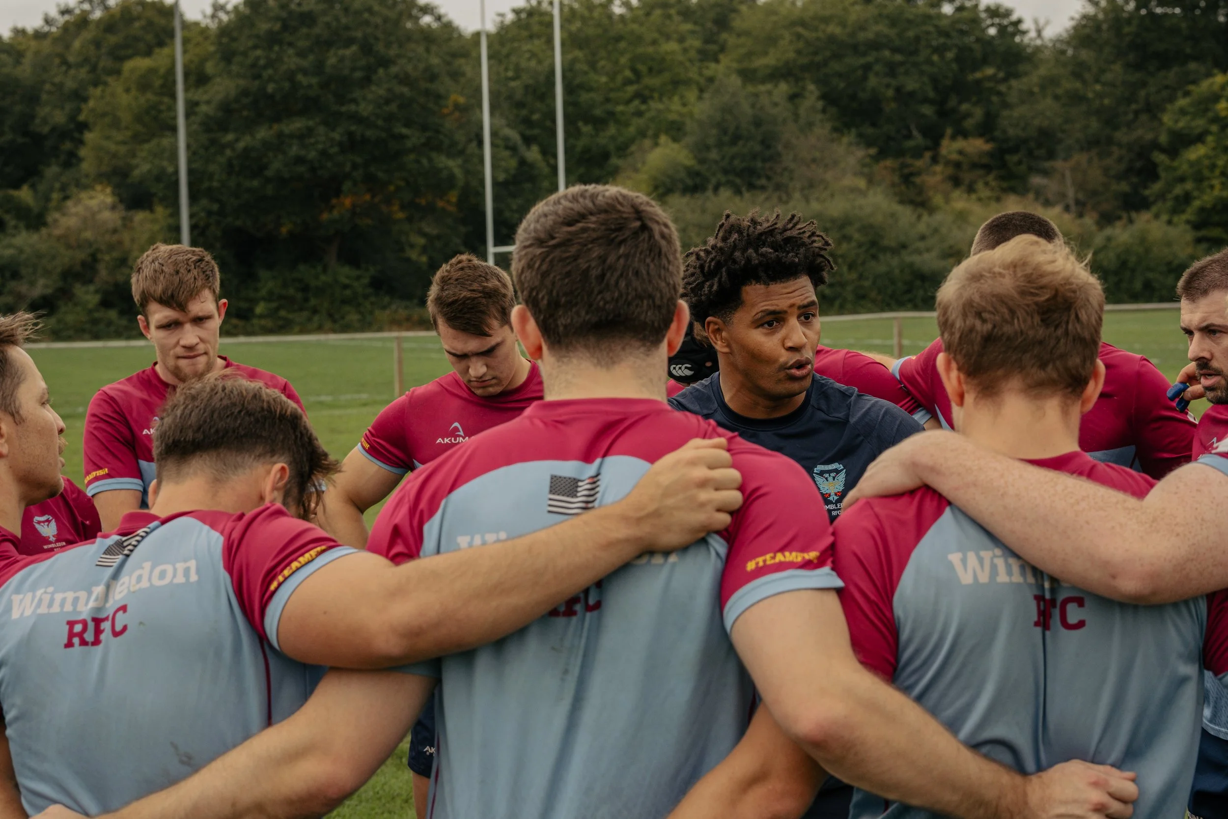 A rugby team huddles together on a field, with players arms around each other's shoulders, listening attentively to a coach or team leader during a practice or game.