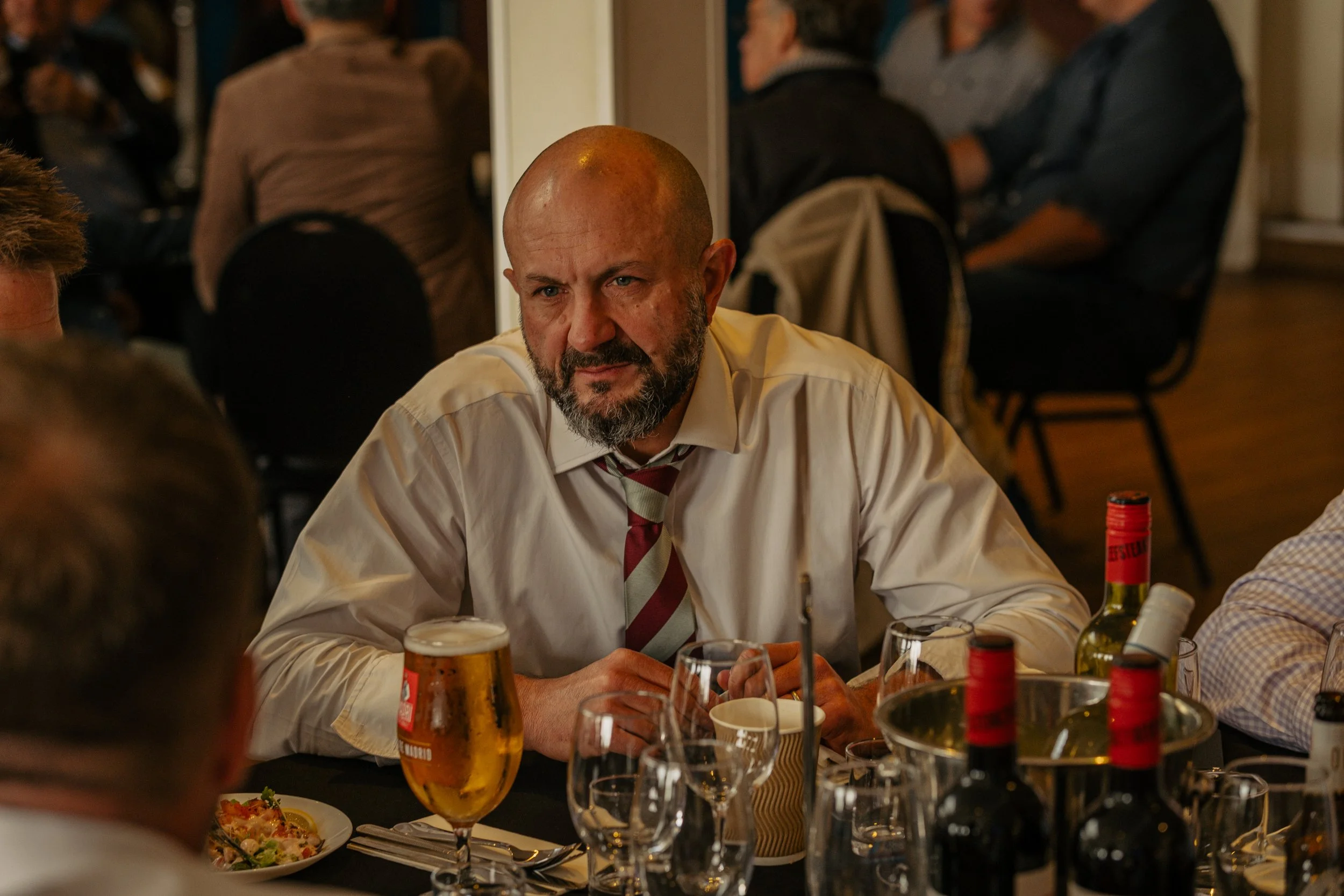 A man with a beard and a bald head, wearing a white shirt and a striped tie, sitting at a table with wine bottles, glasses, and a plate of food, looking intensely at the camera while other people sit in the background.