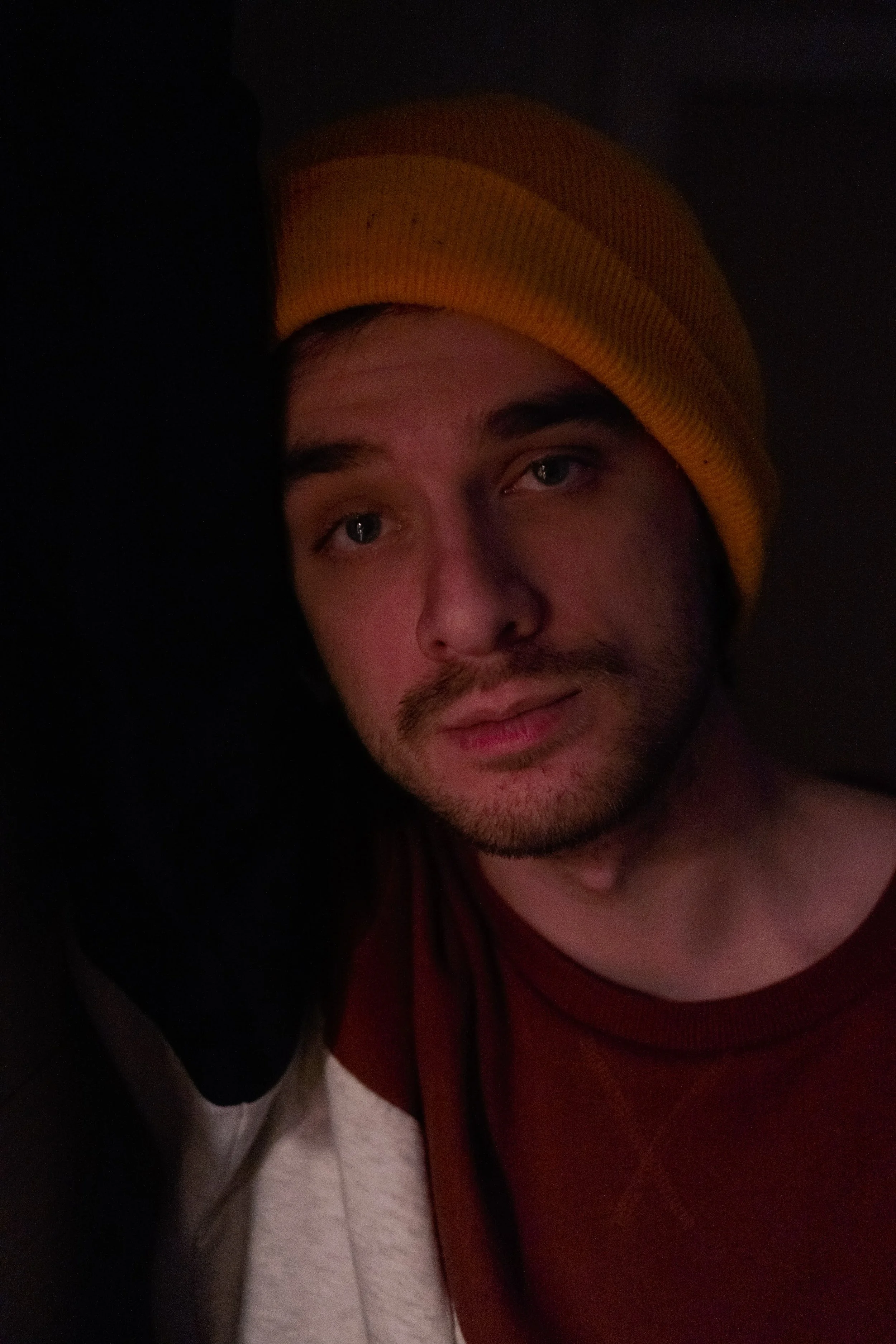 Close-up of a young man with a beard, wearing a yellow beanie, in a dimly lit environment.