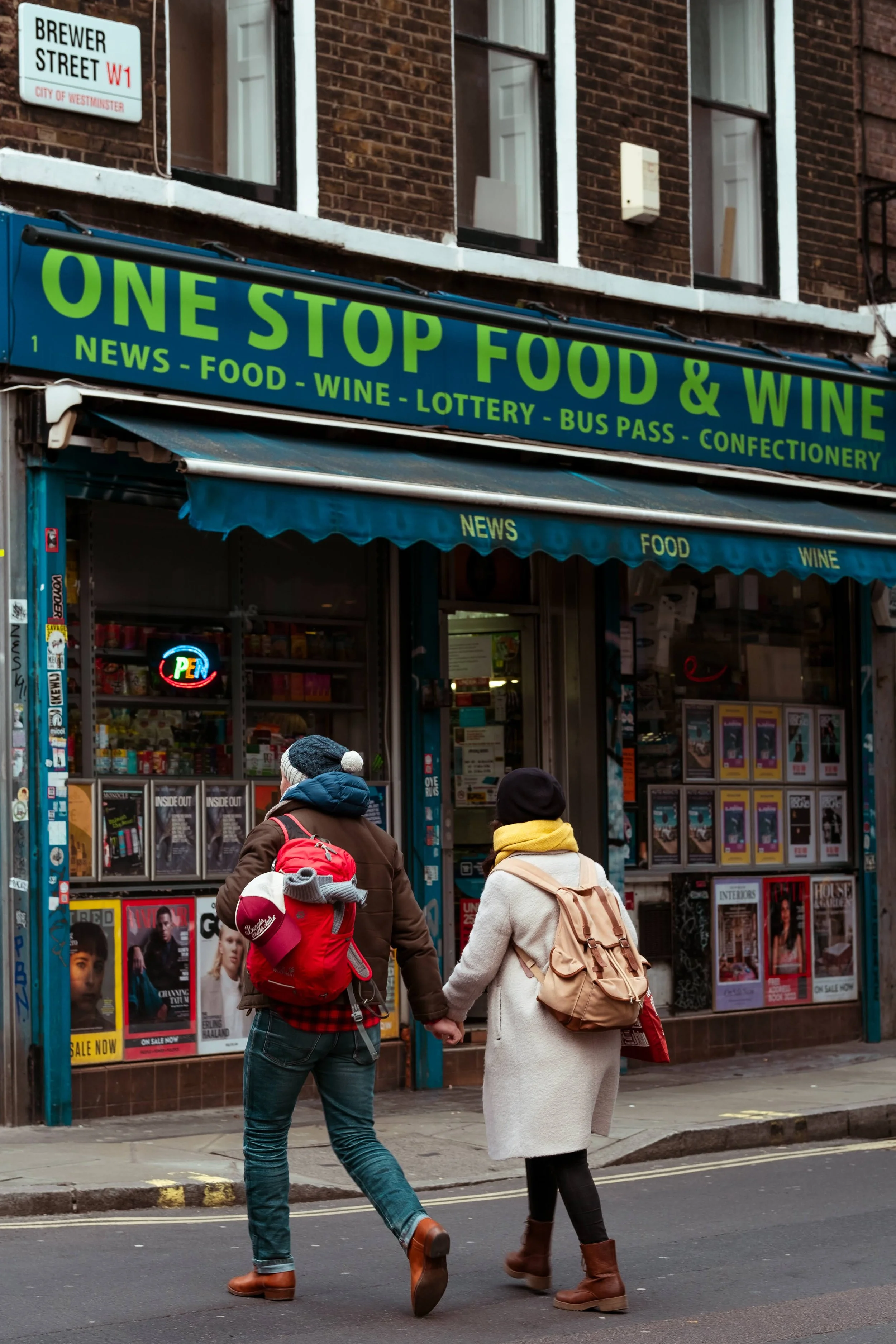 Two people walking hand-in-hand past a storefront named "One Stop Food & Wine" on a city street, with signs indicating it sells news, food, wine, lottery, bus passes, and confectionery.