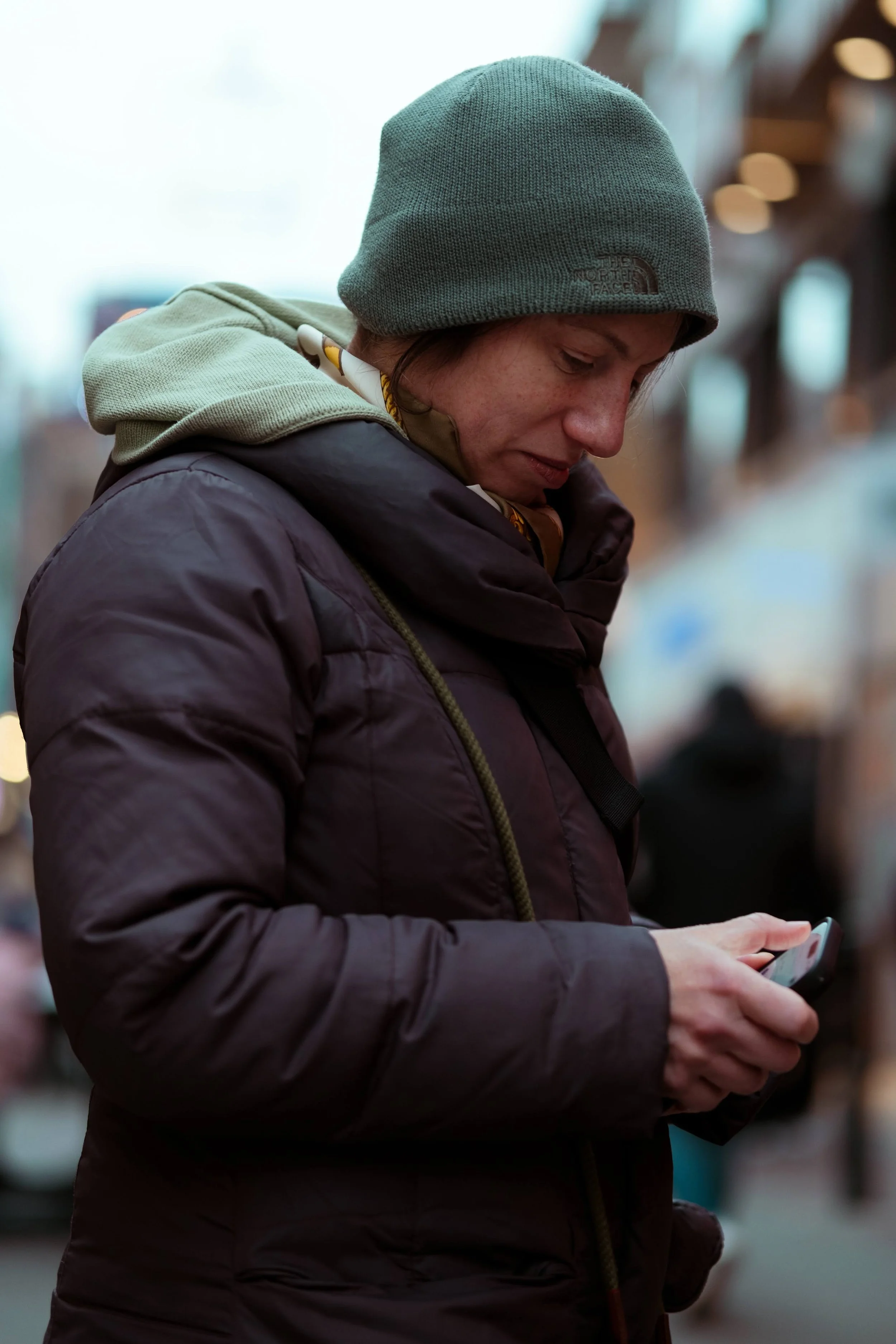 A woman wearing a green beanie, a layered jacket, and a hoodie, looking down while holding and using a smartphone in an indoor setting with blurred background lighting.
