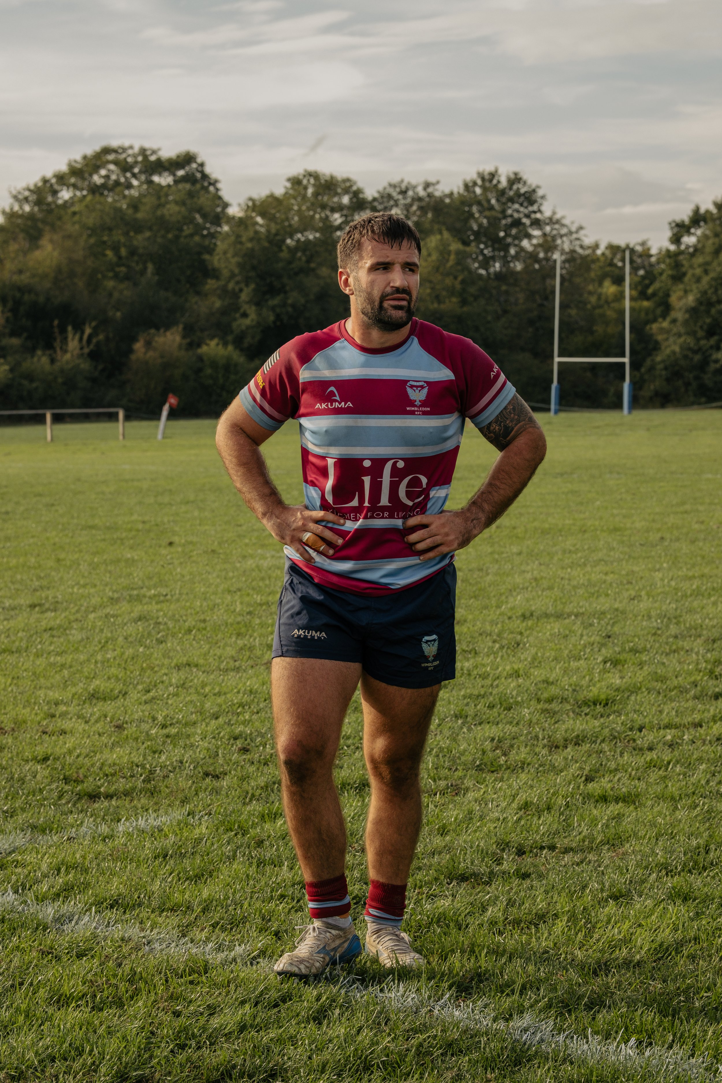 A rugby player standing on the field with hands on hips, wearing a maroon and light blue striped jersey, navy shorts, and rugby cleats, with rugby goalposts and trees in the background.