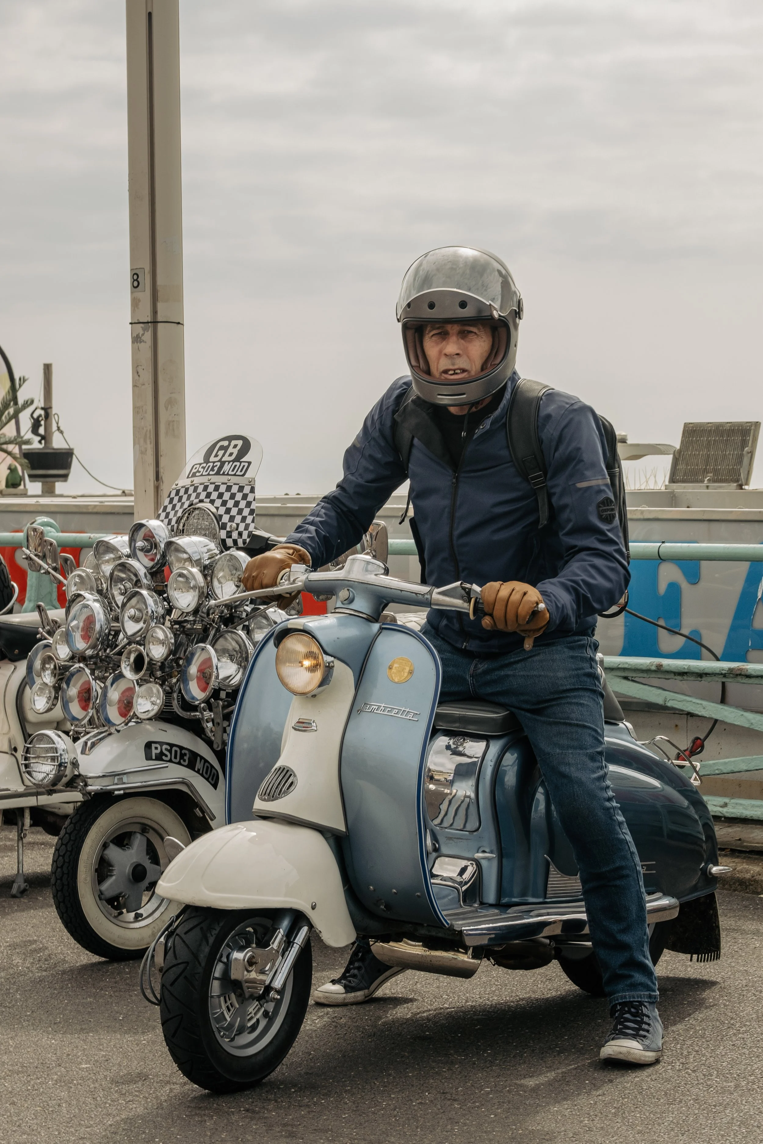 Man in a helmet and jacket sitting on a vintage scooter in a parking lot.