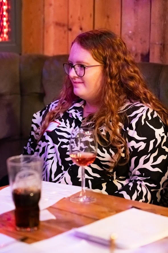 A woman with long red curly hair and glasses sitting at a table with a glass of rosé wine and a glass of cola in a cozy, wooden-paneled restaurant or bar.