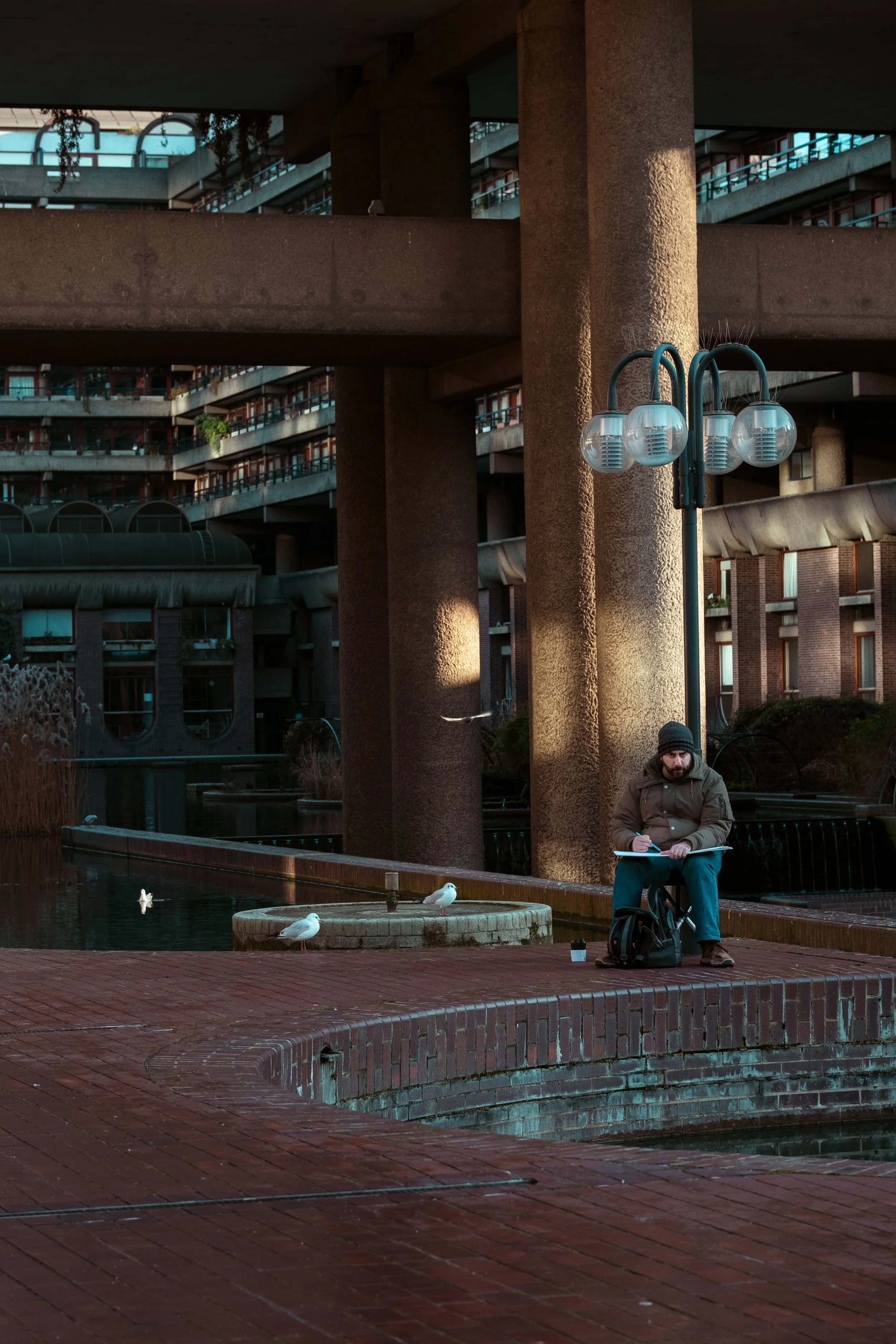 A man wearing a brown jacket, black beanie, and blue jeans, sitting on a brick ledge beside a pond with white seagulls, sketching or writing on a notebook, in an urban park setting with large concrete building structures, a lamp post, and a few birds