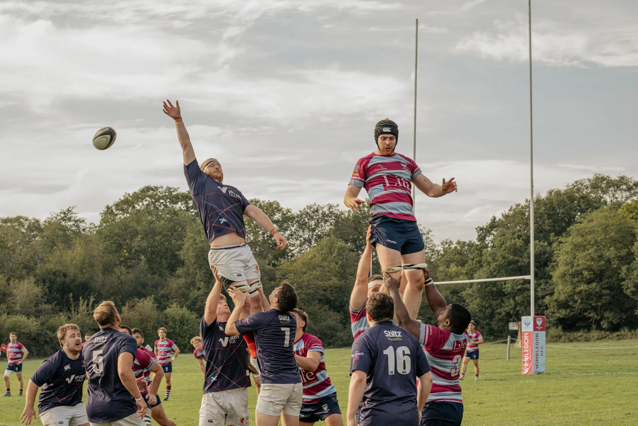 Rugby players in a lineout contest, with two players lifted in the air to catch the rugby ball during a match on a grassy field under a cloudy sky.