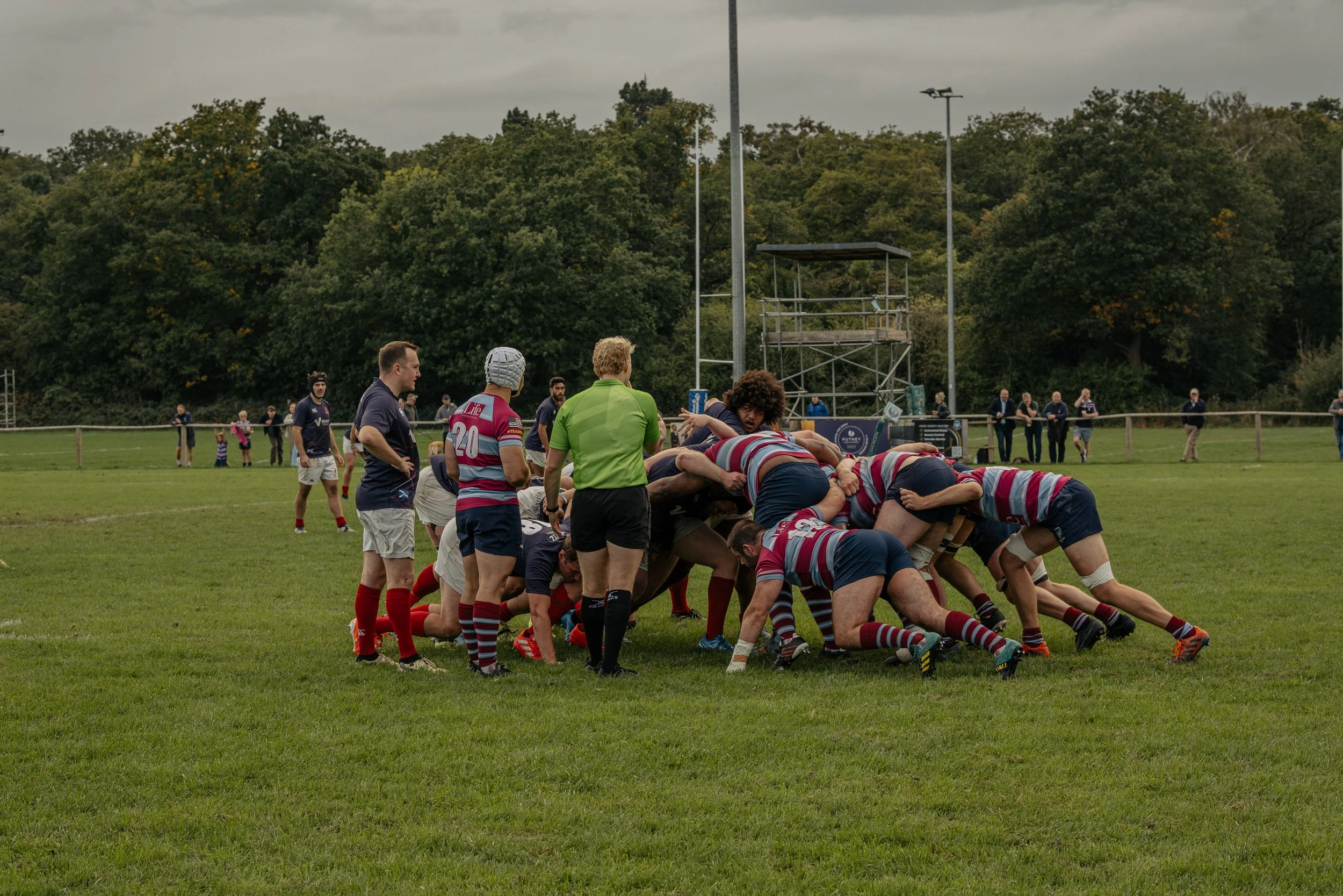 Rugby players engaged in a scrum during a match on a grassy field, with referees and spectators watching in the background under cloudy skies.