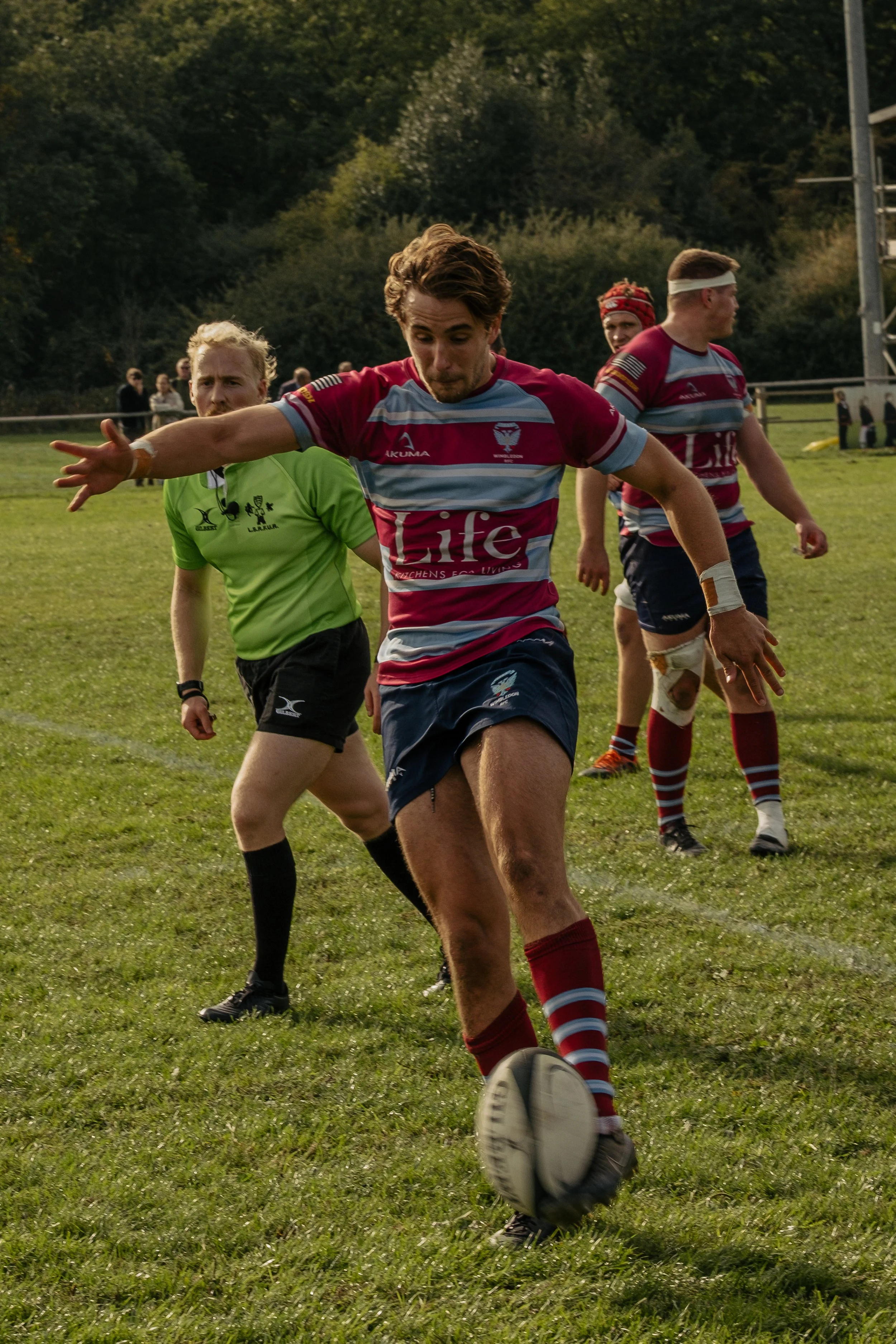 A rugby player in maroon and gray uniform kicking a rugby ball during a game on a grassy field with trees in the background.