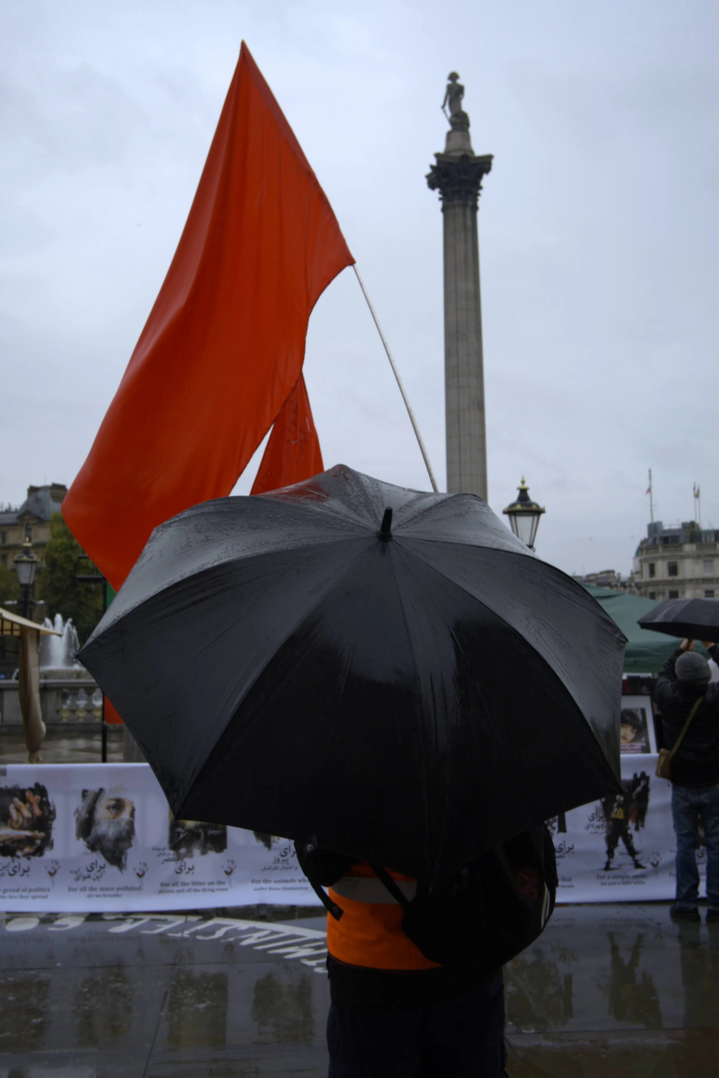 Person holding a black umbrella in front of a protest with an orange flag, and a tall monument in the background, on a rainy day.