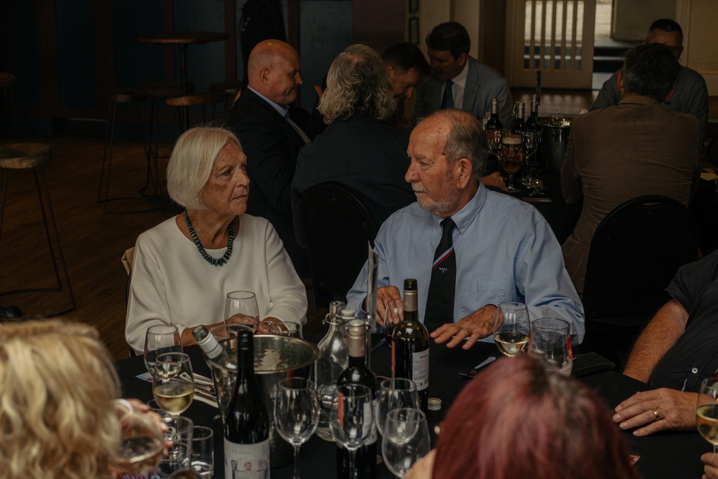 Senior man and woman having a conversation at a dinner party, surrounded by wine bottles and glasses on the table.