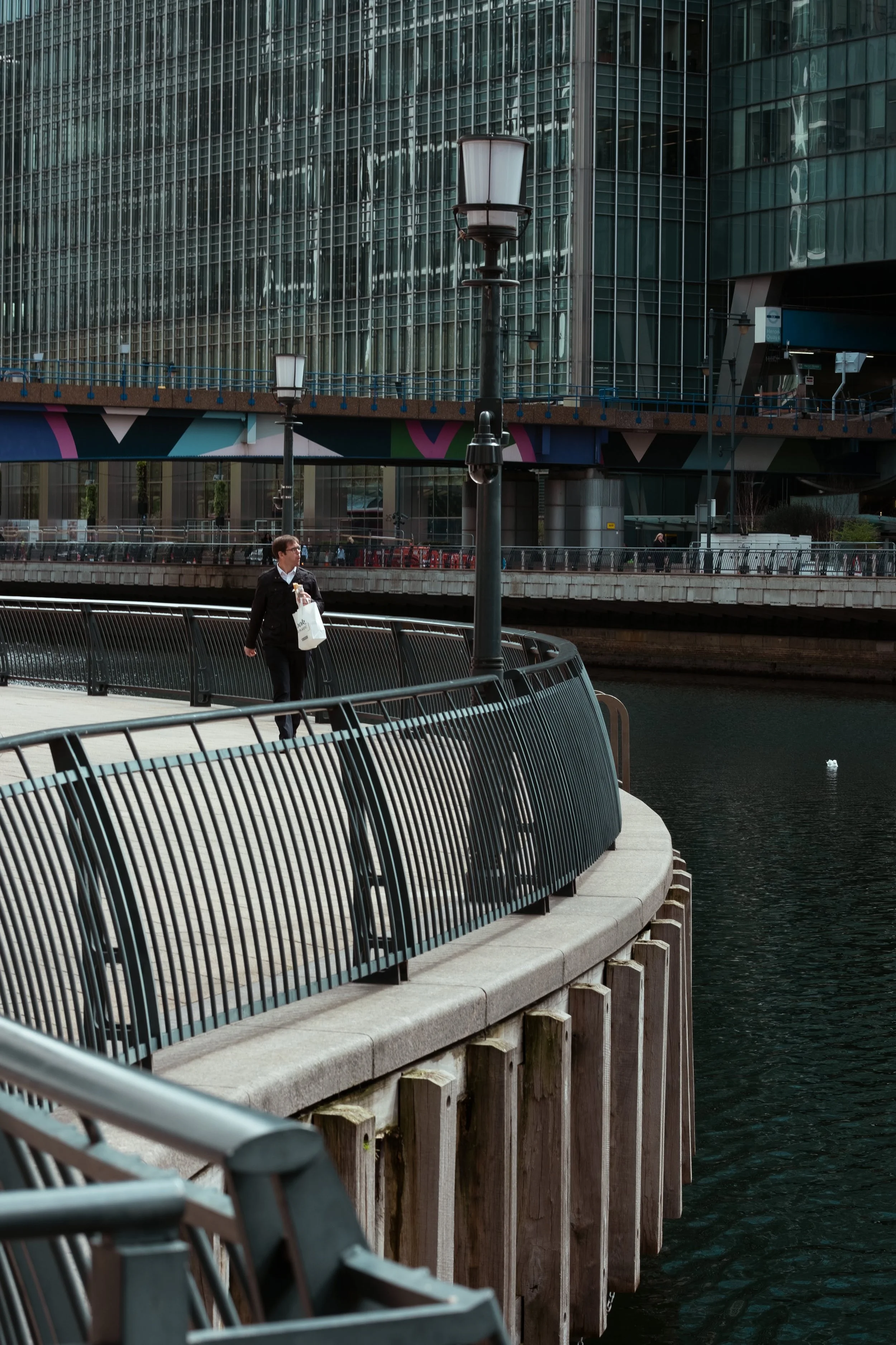 A person walking along a waterfront promenade with a modern glass building in the background, lampposts, and a black metal railing, near a body of water in an urban setting.