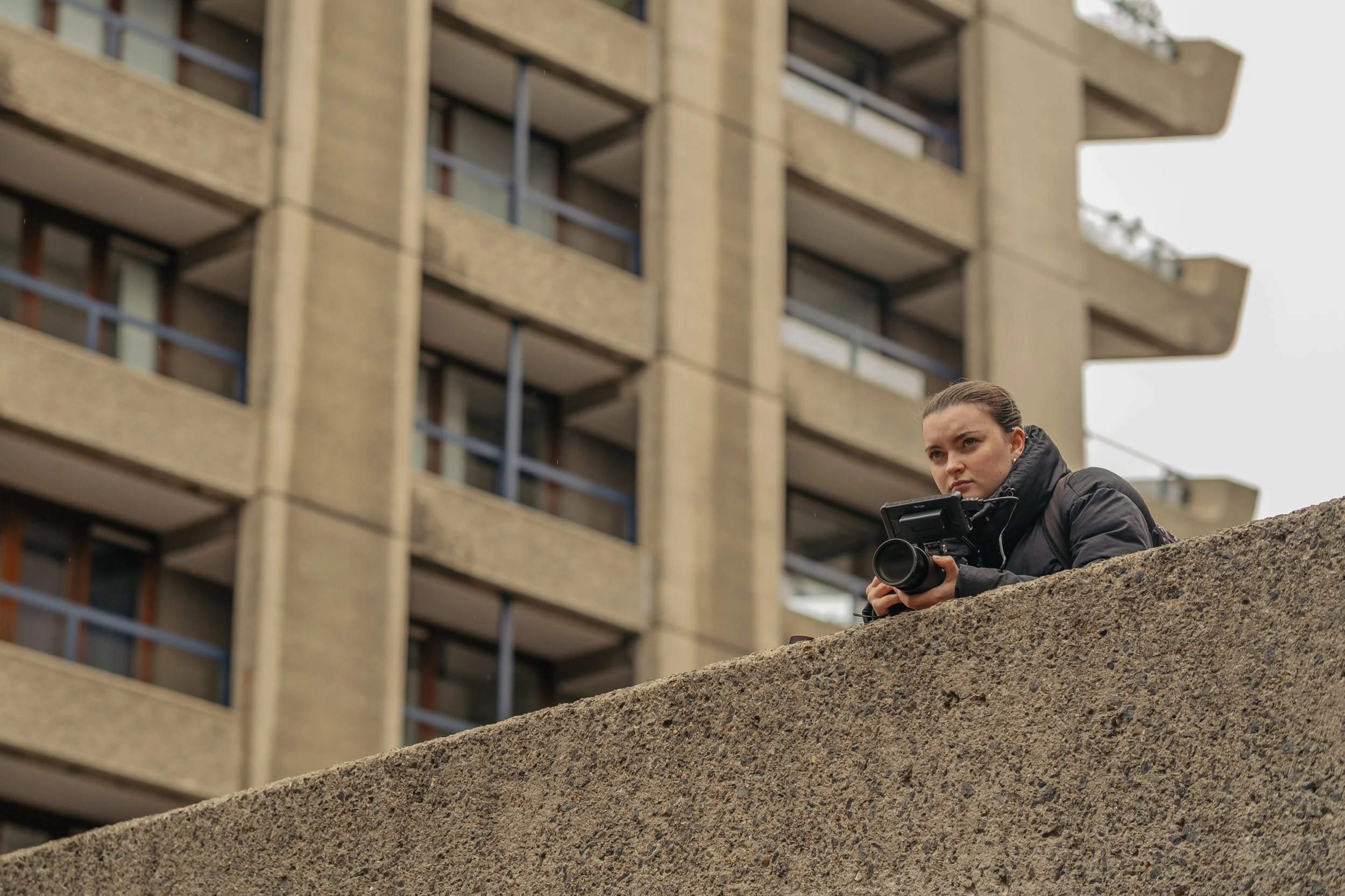 A woman crouching on a concrete ledge, holding a camera, with a modern apartment building in the background.