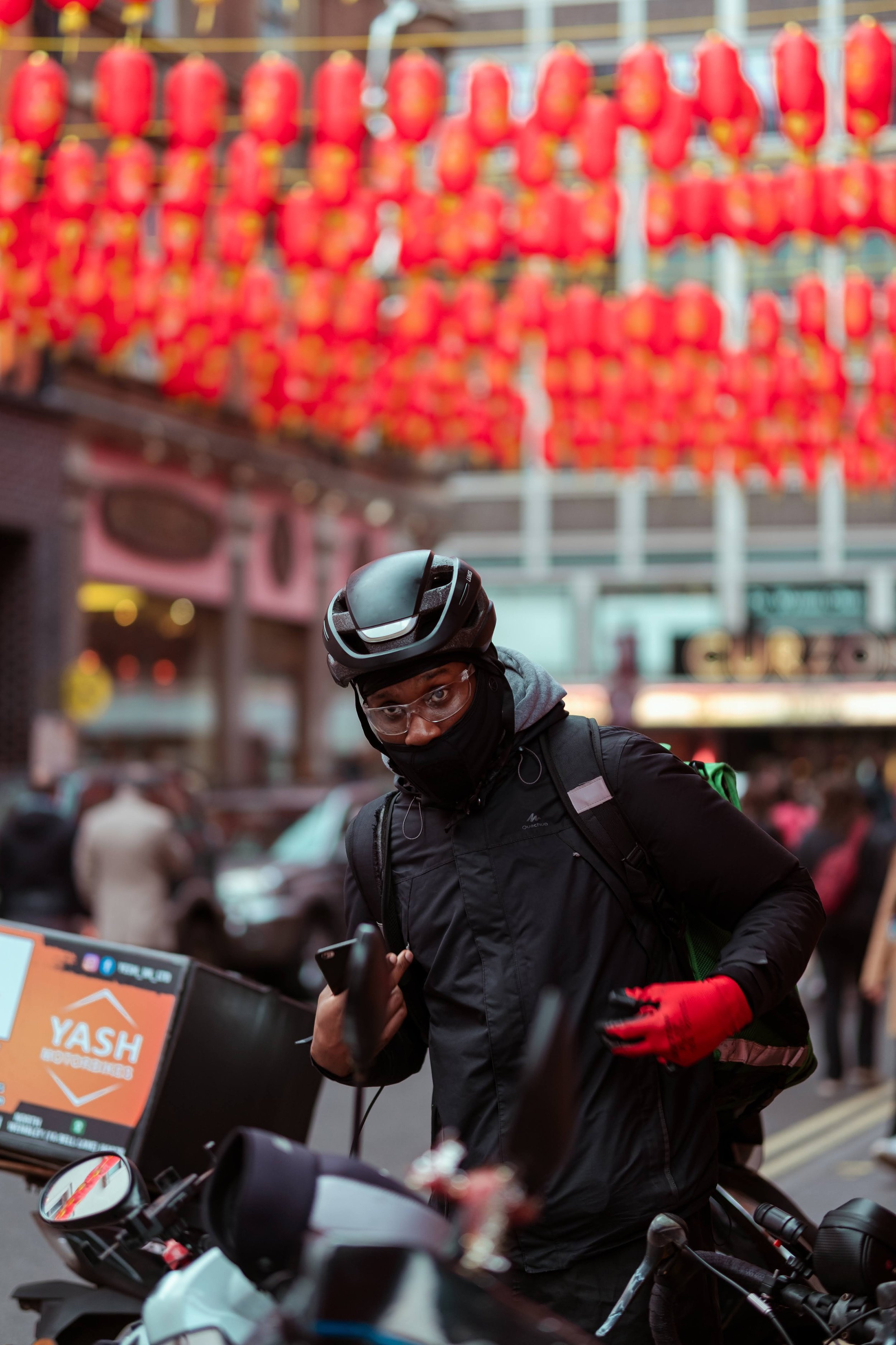 A person wearing a black helmet, glasses, a face mask, a dark jacket, red gloves, and a backpack, standing next to a motorcycle in a busy street with red lanterns hanging overhead in an urban setting.