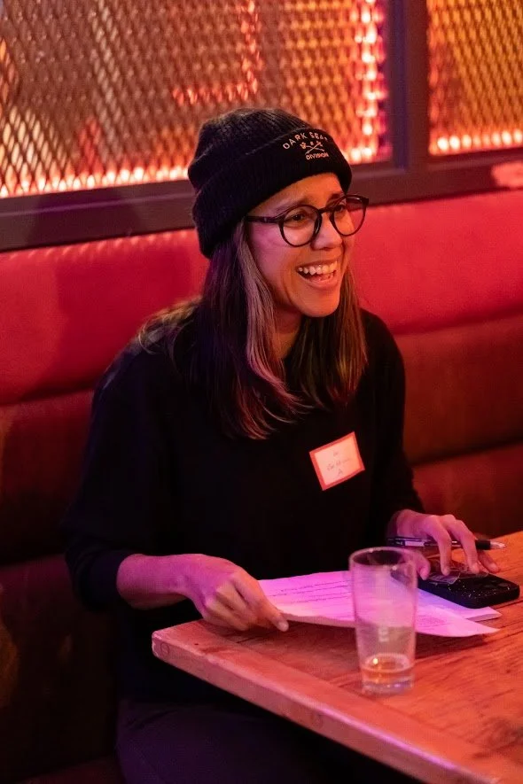 A woman with long hair, glasses, and a black beanie hat, smiling while sitting at a table in a dimly lit setting. She has papers and a remote control in front of her, with a glass of water on the table, and is wearing a black shirt with a name tag.