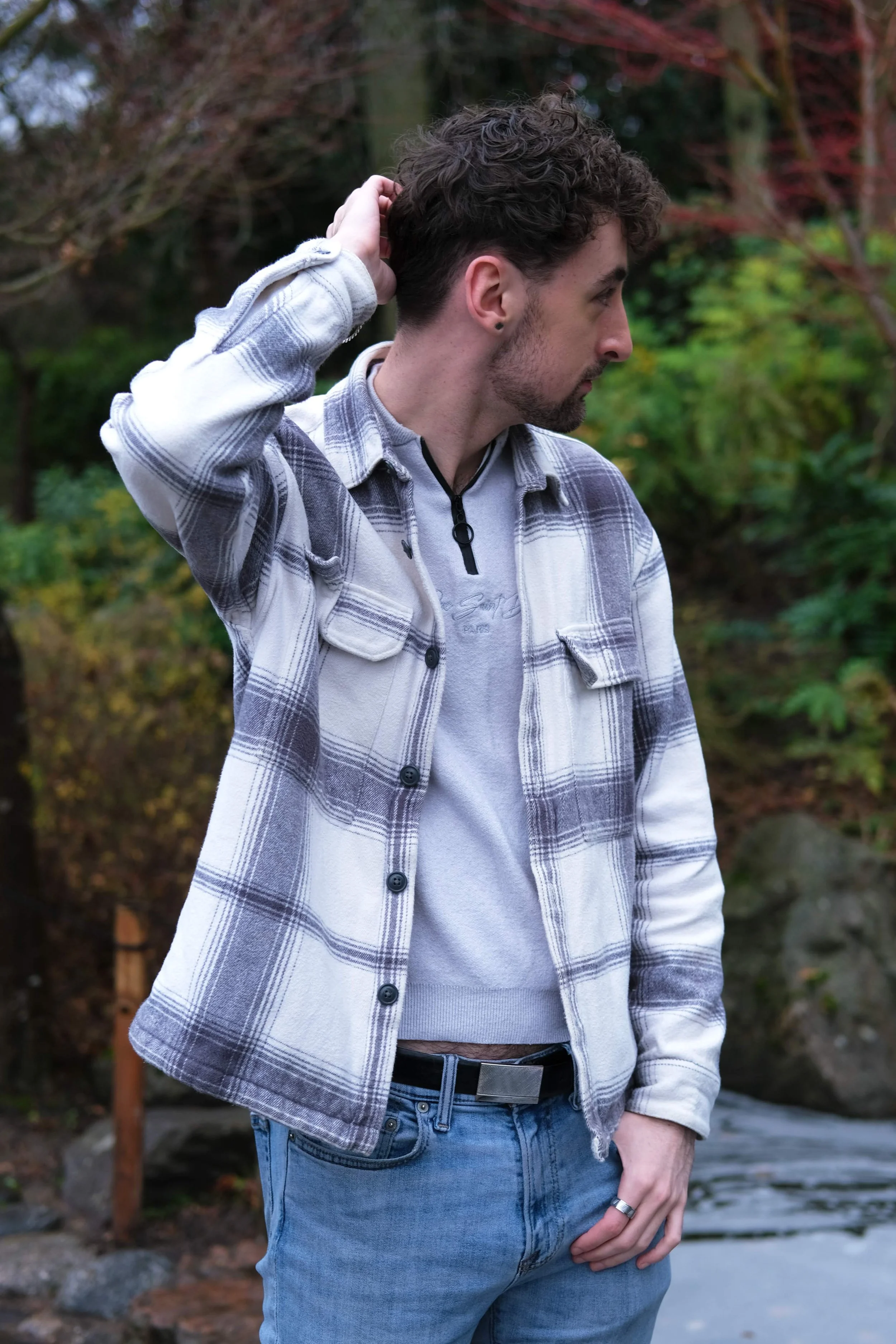 A young man in a plaid shirt and jeans standing outdoors with greenery and rocks in the background, touching the back of his head.