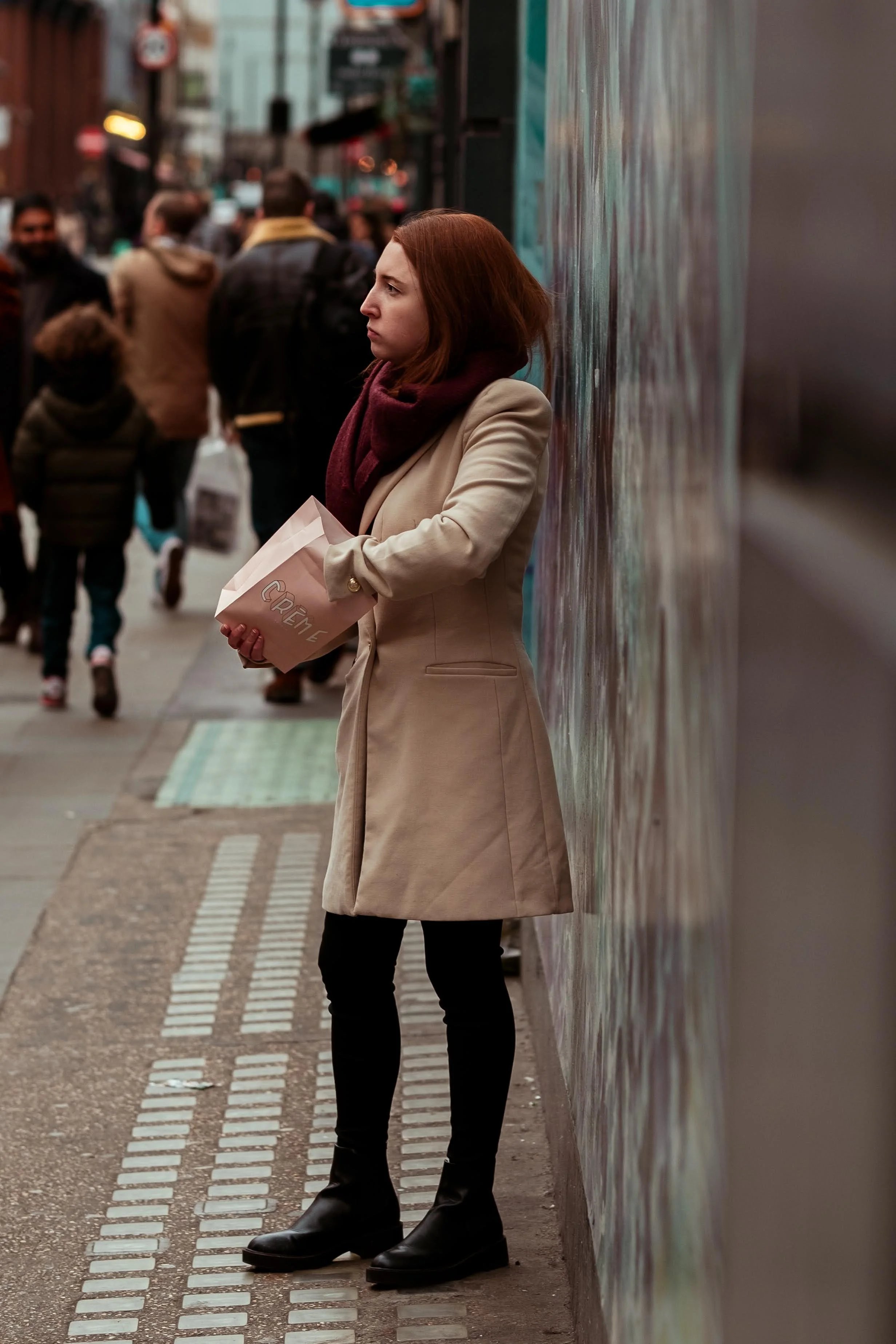 A woman with red hair wearing a beige coat and maroon scarf standing against a colorful graffiti wall on a busy street, holding a white takeout container.