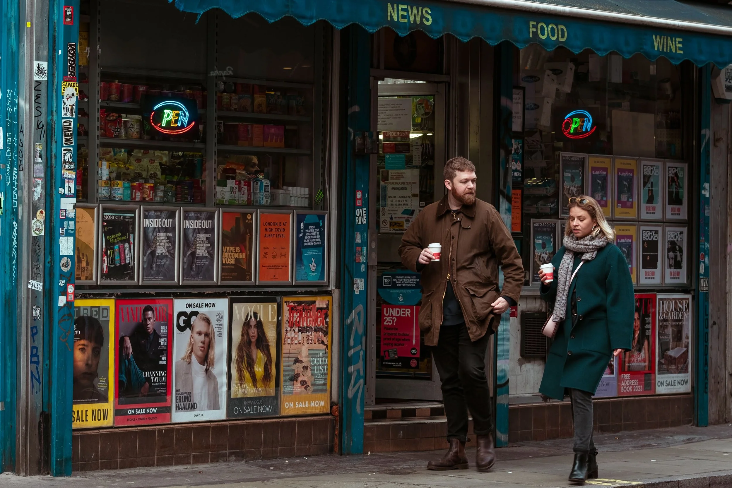 A newsstand shop with magazine racks, neon 'OPEN' signs, and two people standing outside holding coffee cups, engaged in conversation.