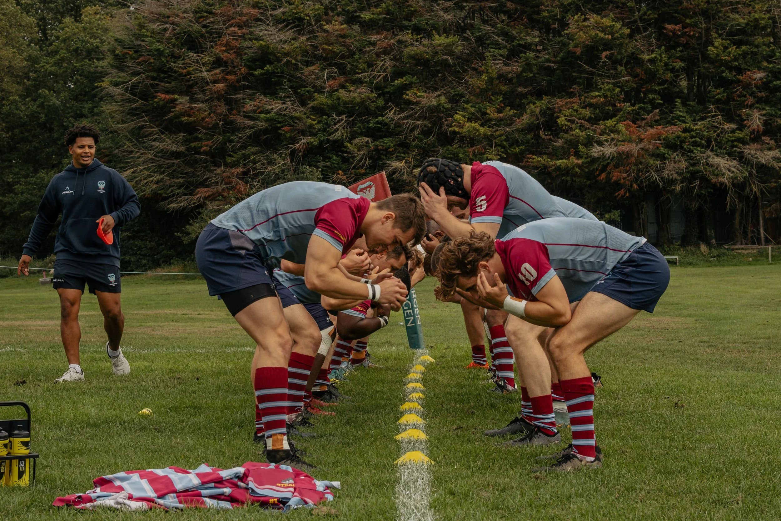Rugby players in grey and red uniforms huddle with their heads down on a field, practicing a scrum or preparing for a play, with a coach observing in the background.