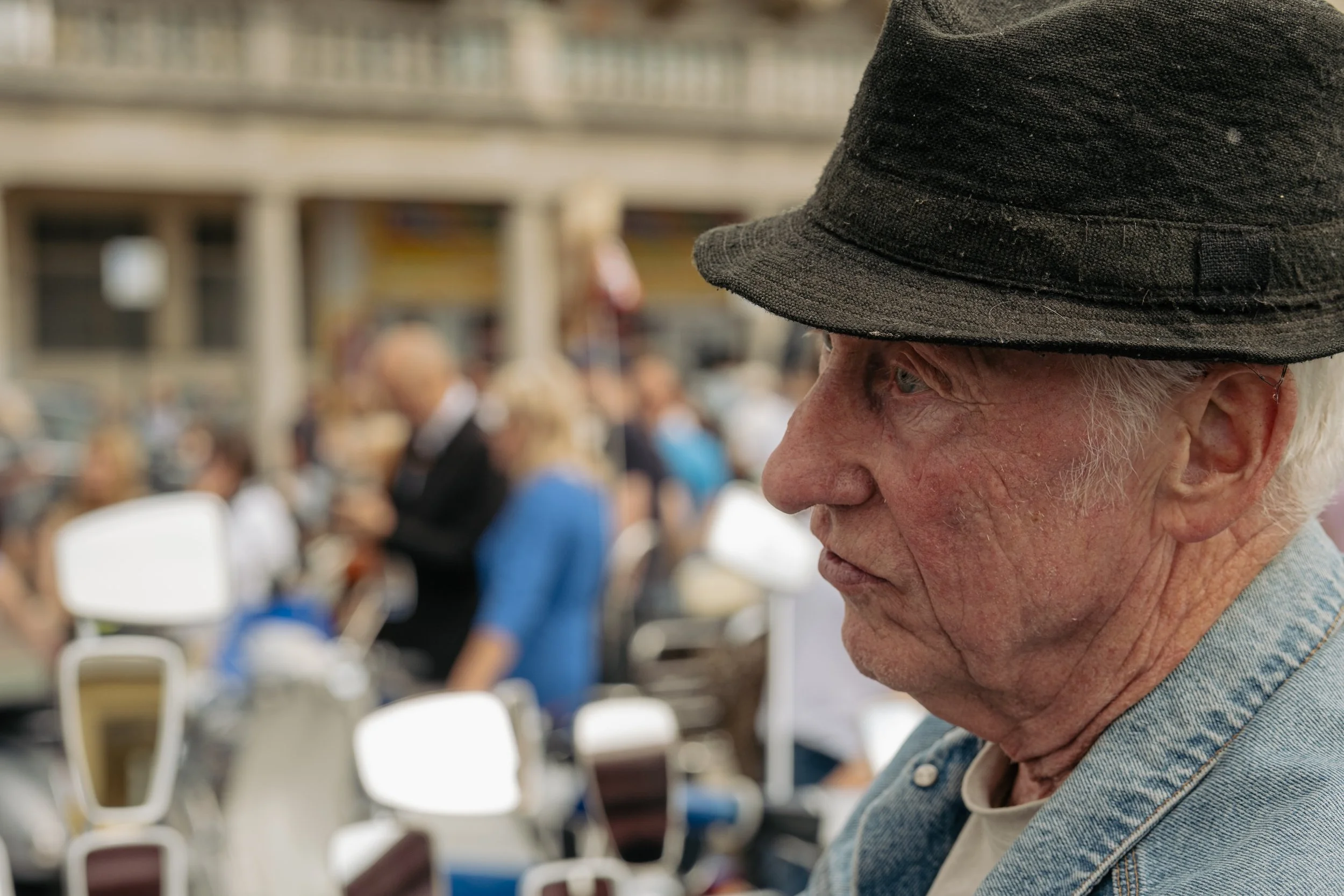 Close-up of an elderly man wearing a black hat and denim jacket, at an outdoor event with a blurred crowd in the background.