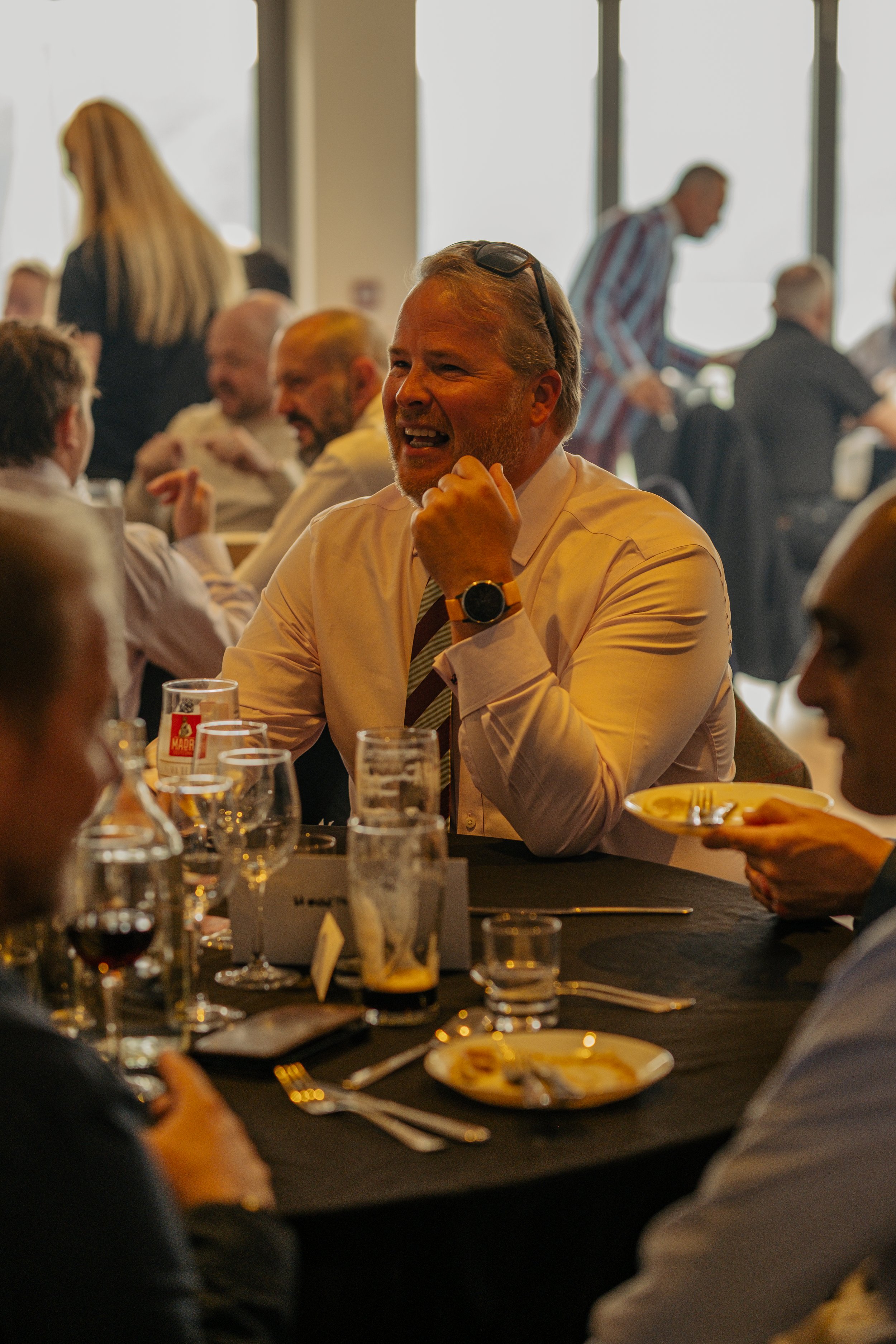 A group of well-dressed people at a formal event or dinner, with one man smiling and appearing to speak at a table with drinks and plates of food, in a room with large windows and natural light.