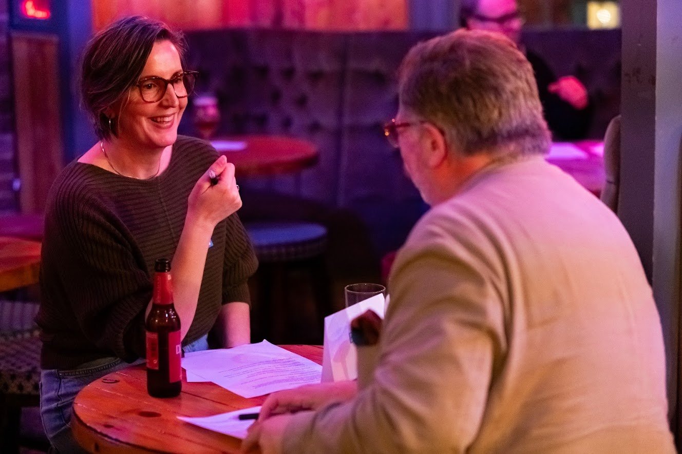 A woman with short brown hair and glasses, smiling and holding a pen, is talking to a man with glasses and gray hair, who is looking at her. They are sitting at a round wooden table in a dimly lit bar or restaurant, with a beer and papers on the tabl