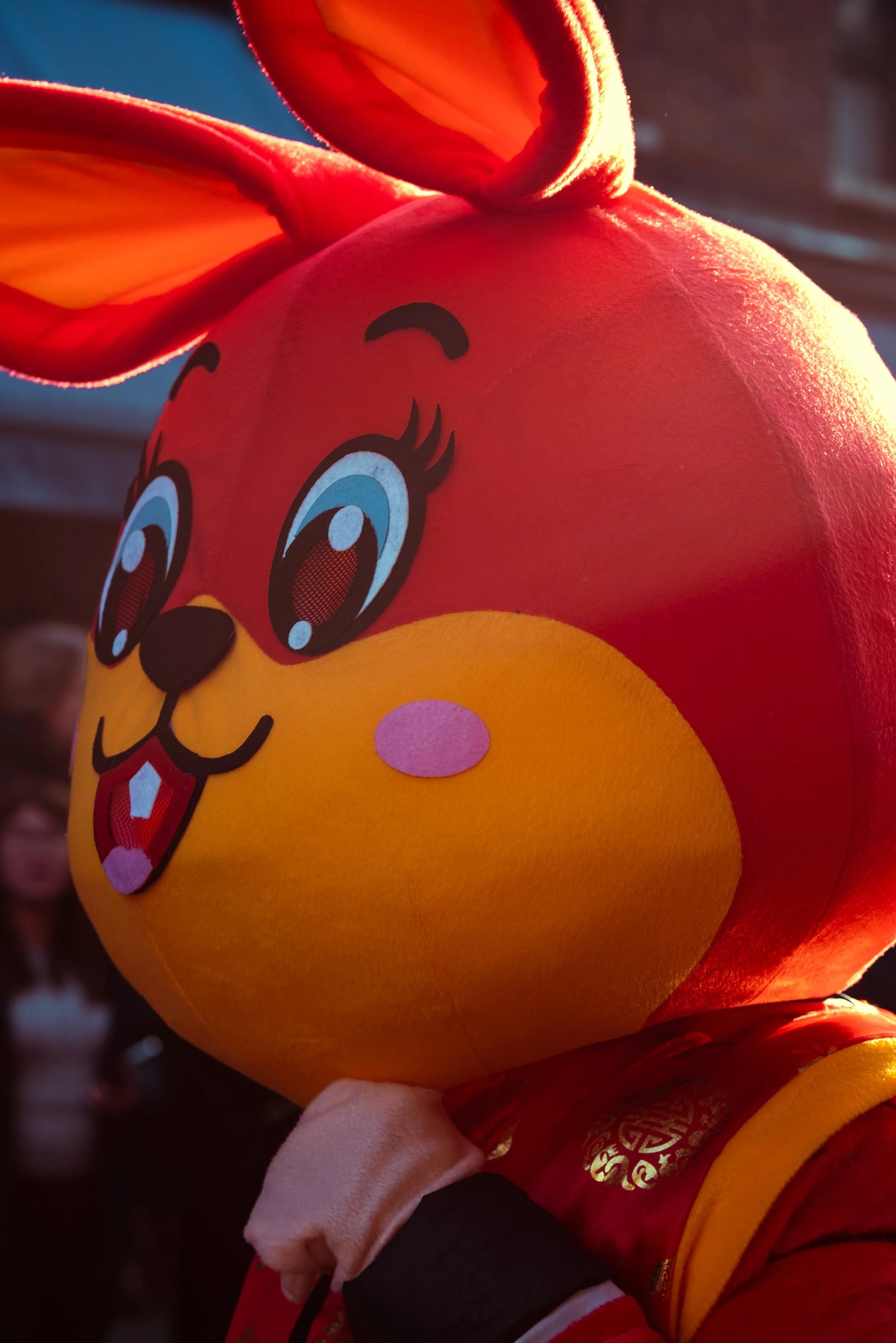 Person wearing a large cartoon rabbit mascot head with big eyes, pink cheeks, and pink tongue, in a red and yellow costume, illuminated by sunlight.