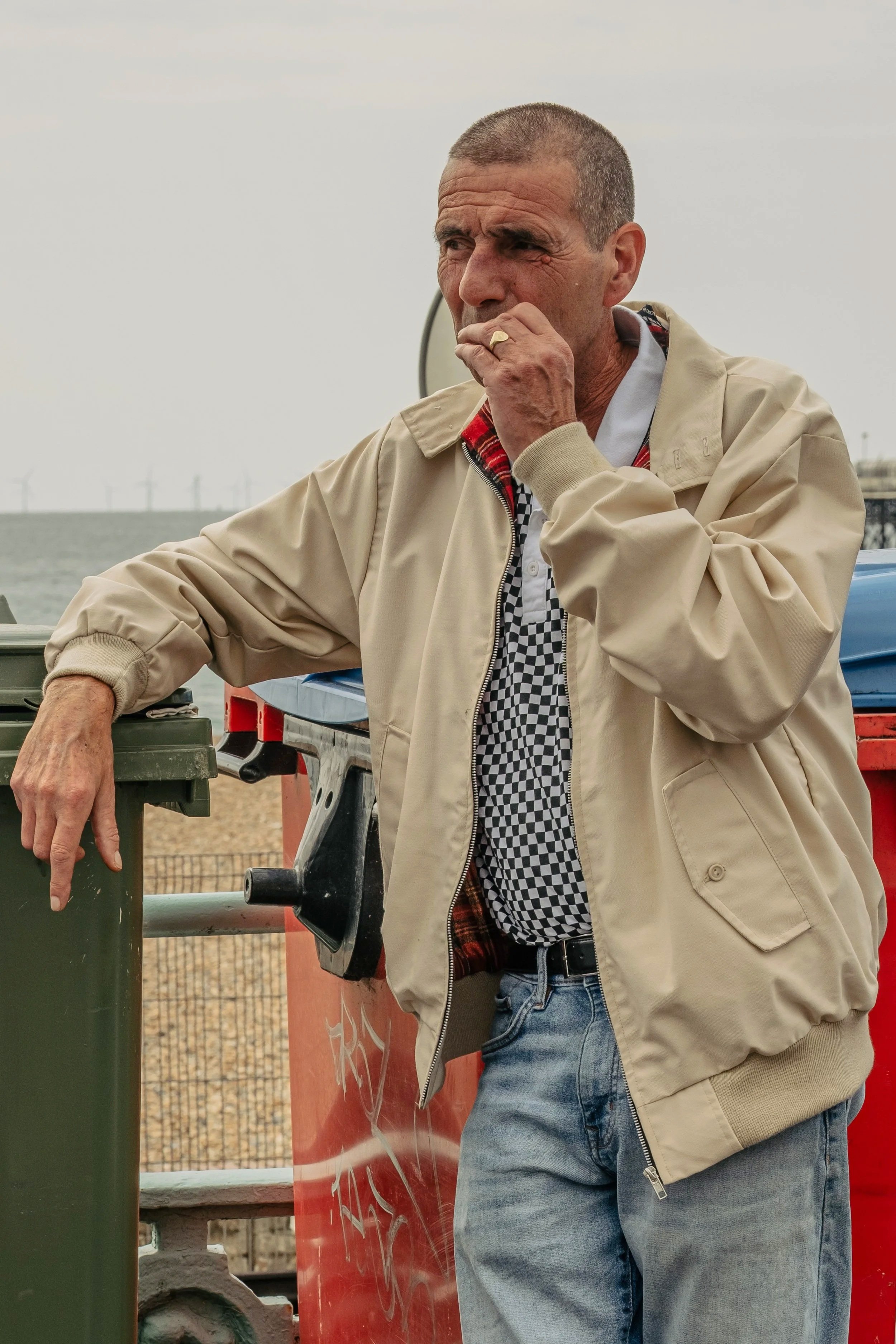 Man in beige jacket and blue jeans standing outdoors near a trash can, with an overcast sky and a distant view of a pier or bridge in the background.