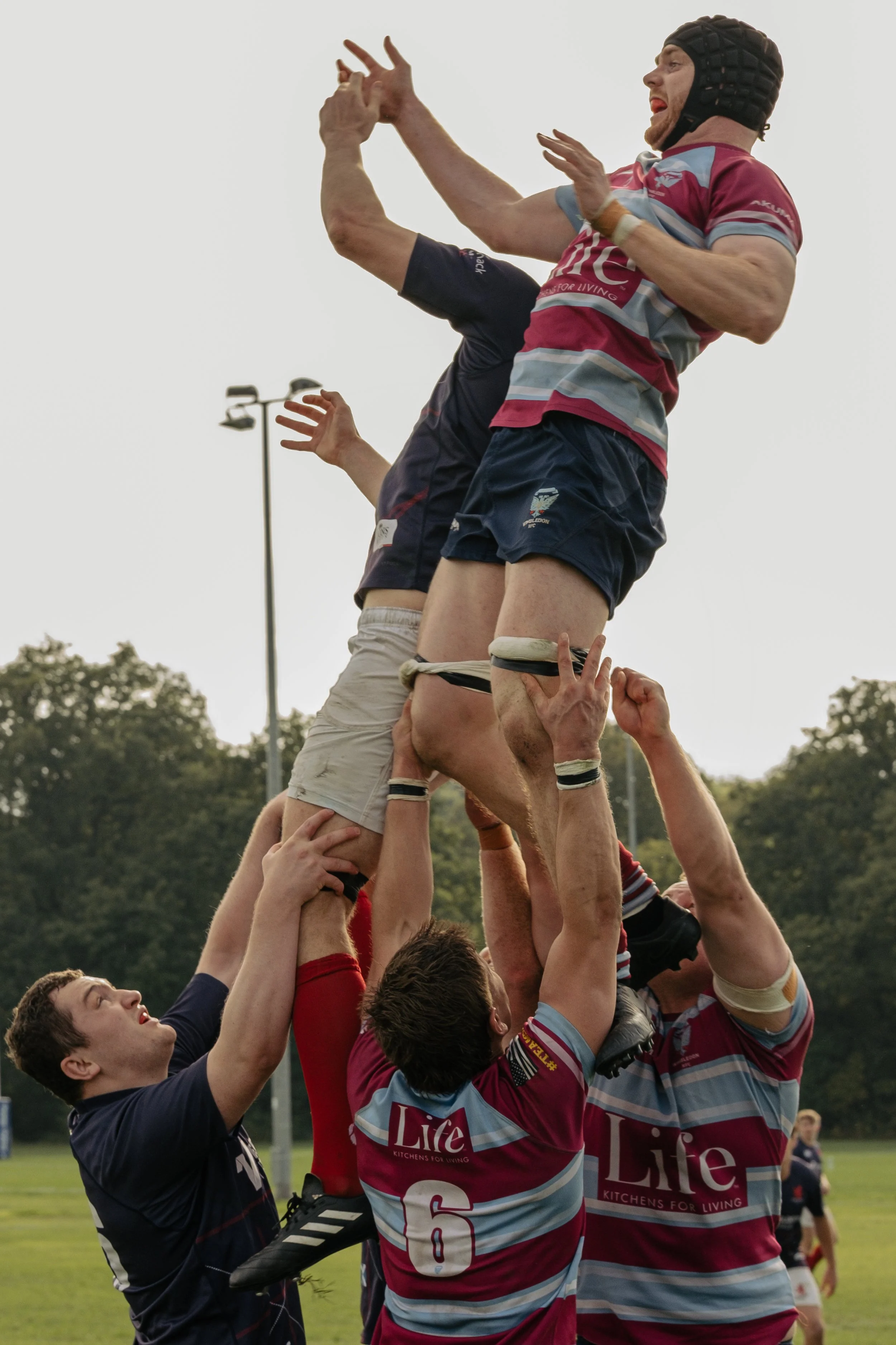 A rugby crowd cheer as players lift a teammate into the air during a lineout on a grassy field.